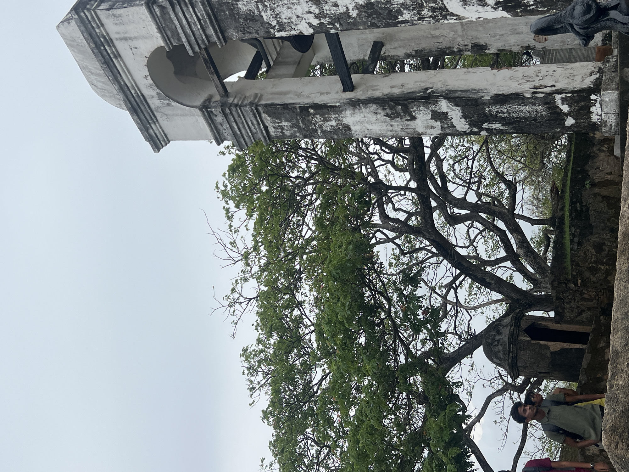 In Galle, Sri Lanka, a large, weathered bell tower stands to the right of the frame, its white facade stained with dark patches of age and grime. Arches reveal dark bells suspended within. To the left, a lush, sprawling tree with a thick, gnarled trunk dominates the middle ground, its branches reaching across the sky. Behind the tree, a small, stone turret-like structure with a dark opening is visible. In the foreground, two young men are walking on a stone pathway. One, wearing a green polo shirt and backpack, looks towards the left. Another person, partially visible, wears a red shirt. The sky above is overcast and a pale grey.