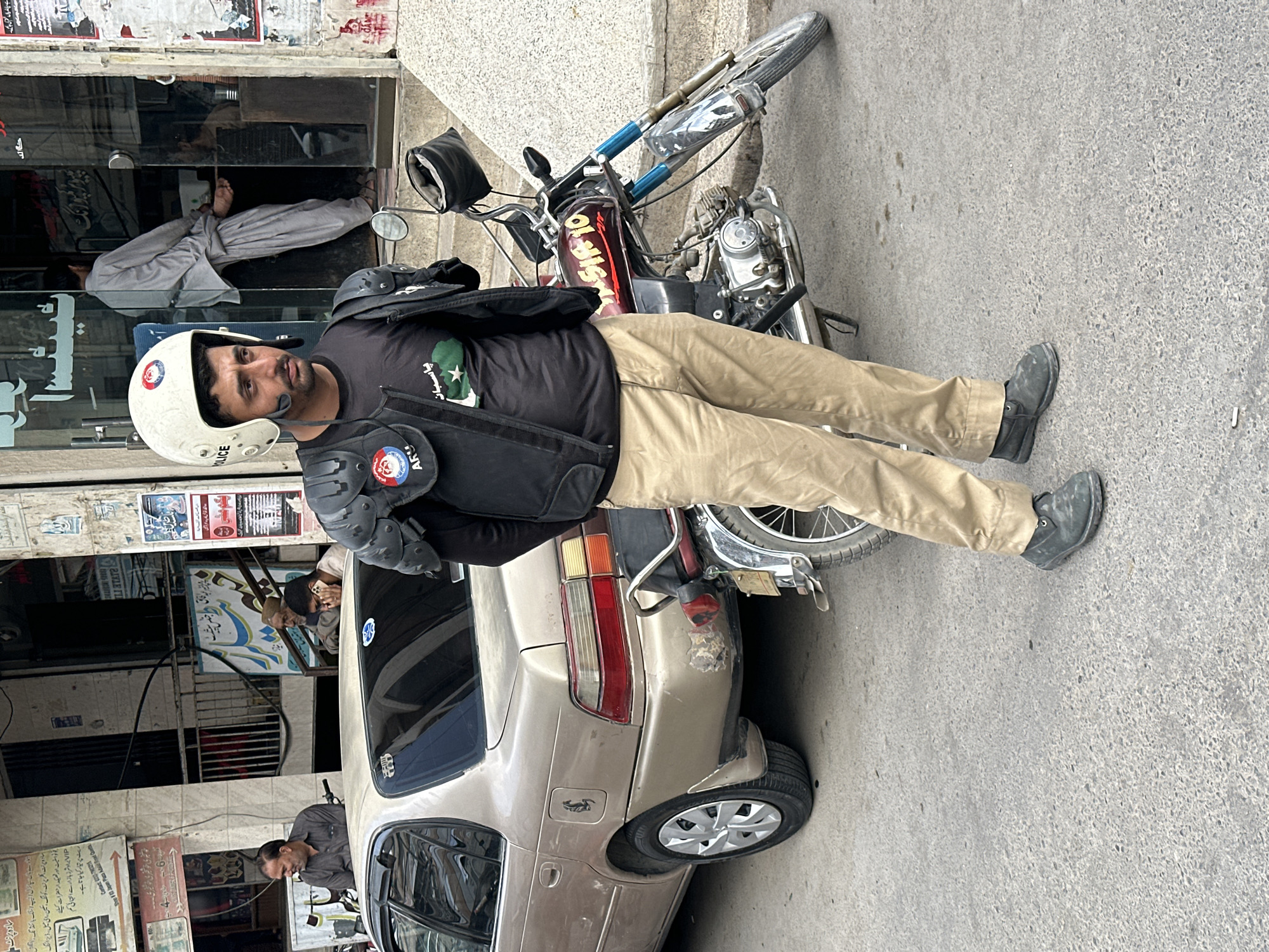 A police officer in uniform, including a helmet and protective gear, is standing on a city street. He is wearing a black shirt with a Pakistan flag emblem and khaki pants. A motorcycle is parked behind him, leaning against a parked gold-colored car. Several people are visible in the background, observing from doorways and windows. The location appears to be a commercial area within Quetta City Tehsil, Pakistan, judging by the text on storefronts and the style of dress of people in the background.