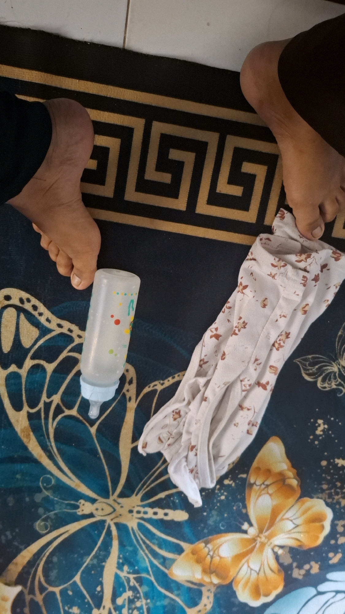 The image depicts a close-up, overhead view of a dark blue rug with a butterfly pattern. Two bare feet are visible on the rug. On the left, a left foot with visible toes and toenails rests on the rug. On the right, a right foot, also with visible toes and toenails, is positioned slightly higher on the rug. Between the two feet, a clear baby bottle with a blue nipple and multicolored polka dots is lying on its side. To the right of the bottle and foot, a light-colored piece of fabric with a floral pattern is crumpled. The rug's pattern includes a large, stylized golden butterfly in the foreground and a Greek key border in black and gold at the top. The overall scene suggests a domestic setting. The specific location in Bandar Lampung, Indonesia, cannot be determined from the image.