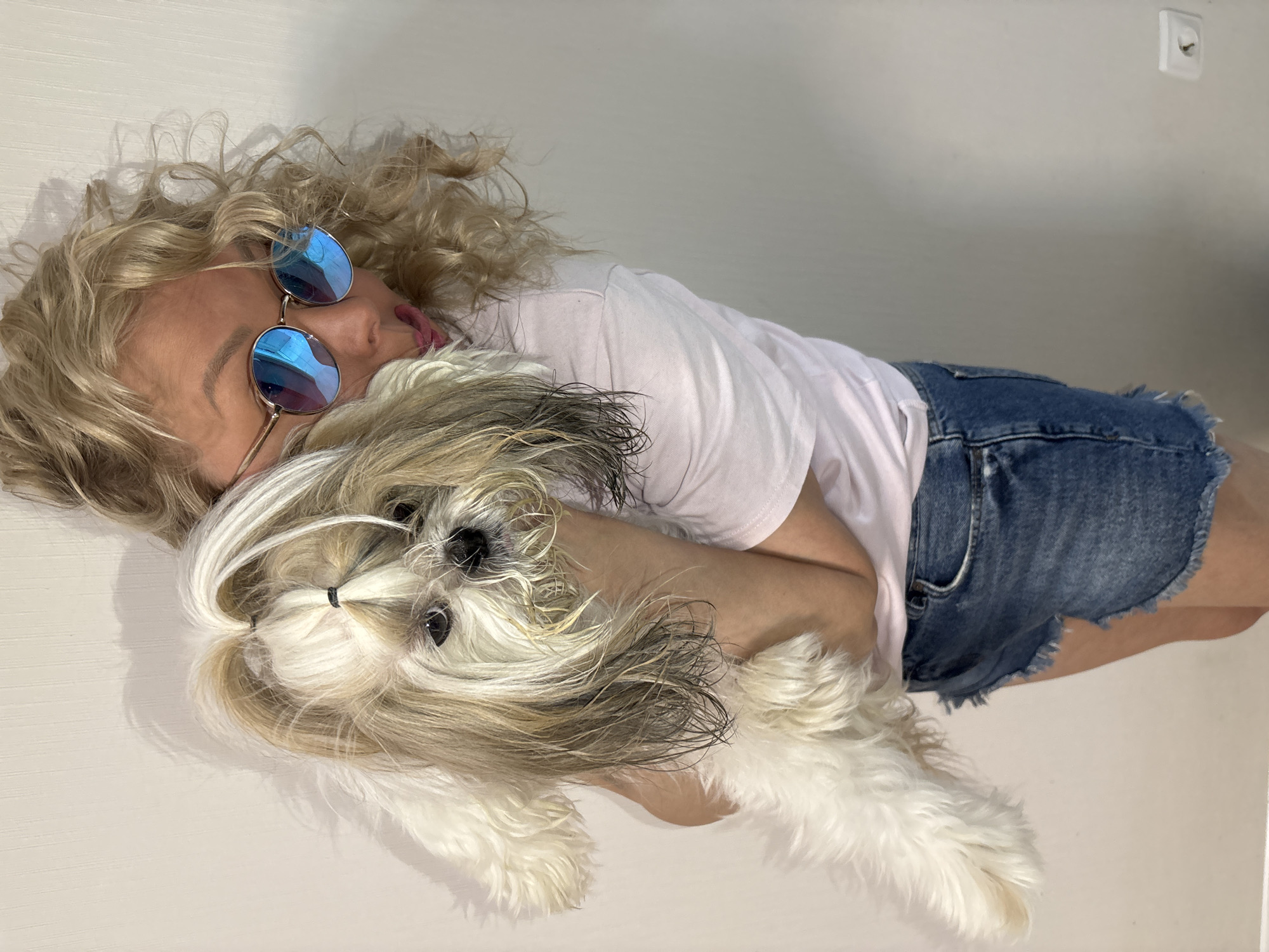 A blonde woman wearing blue sunglasses and a white t-shirt is lying on her side, embracing a Shih Tzu dog with white and brown fur. The woman is also wearing blue denim shorts. They are lying on a white surface against a white wall. An electrical outlet is visible on the wall. It is not possible to determine if this image was taken in Izhevsk, Russia.