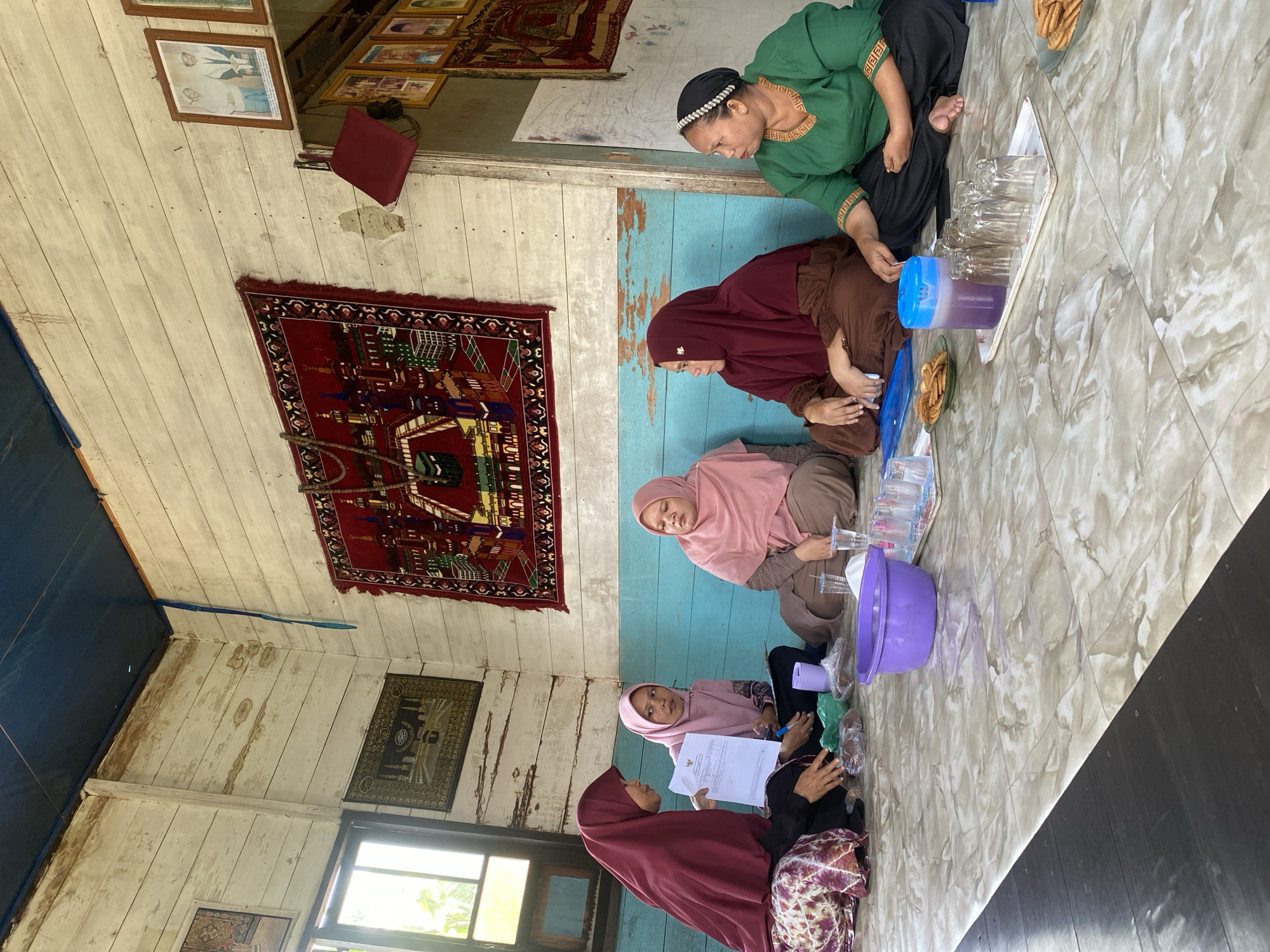 In Bawahan Selan, Indonesia, five women are gathered inside a rustic wooden building, seated on a tiled floor. The room features weathered white wooden walls, a dark blue ceiling, and a turquoise-painted section of the wall. Decorative rugs depicting Islamic scenes hang on the walls. The women, all wearing hijabs or head coverings, appear to be engaged in a meeting or discussion. One woman on the left, wearing a maroon hijab, holds a document and is speaking. The others are listening or observing. On the floor in front of them are various items, including a purple basin, packets of food, cookies on a plate, a jug of purple liquid, and a tray with several glasses. The natural light entering from a window suggests it is daytime. The overall impression is a casual gathering in a simple, traditional Indonesian setting.