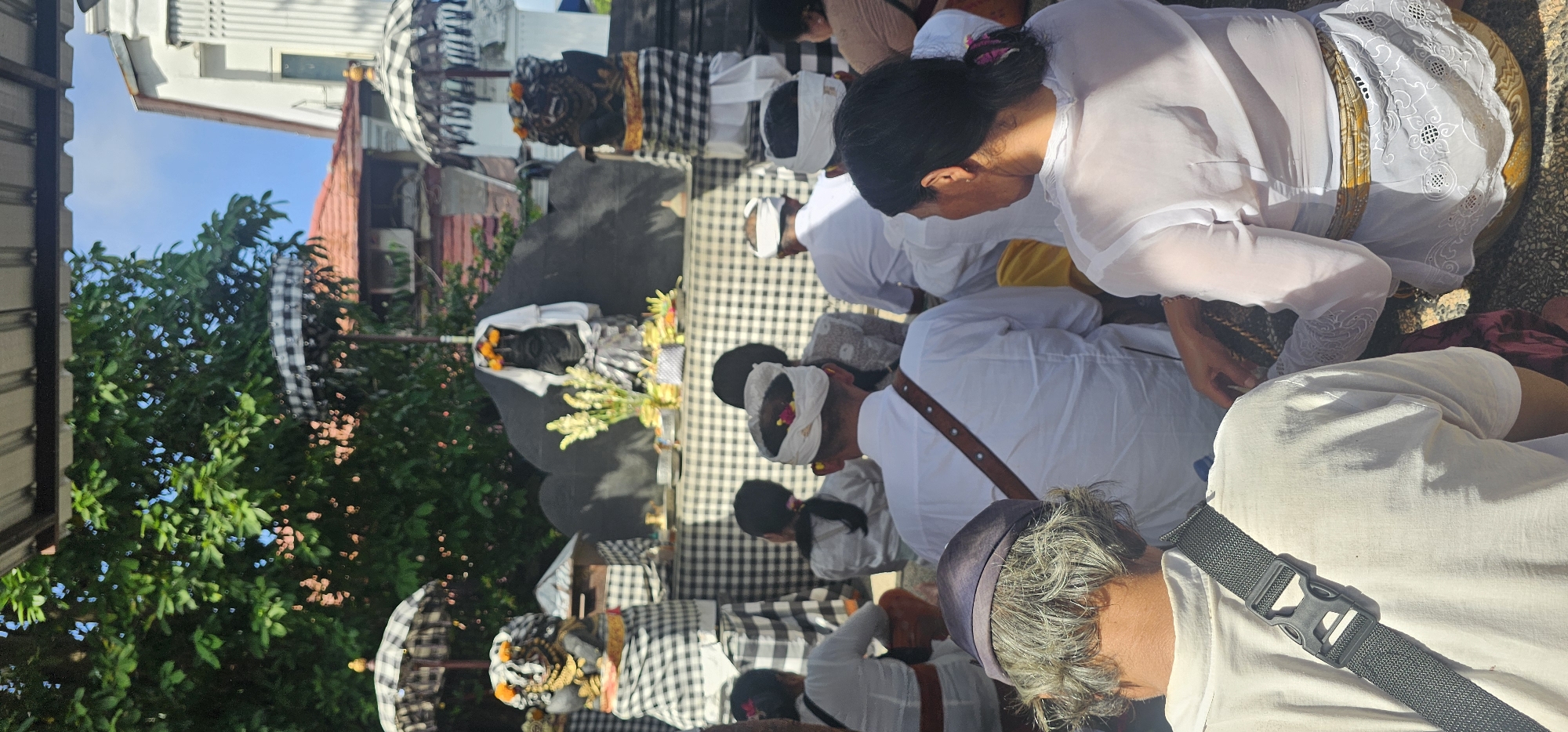 This image captures a religious ceremony taking place in Kuta, Indonesia. Several people are kneeling on a checkered surface, dressed in white traditional attire. One woman in the foreground is wearing an ornate white lace outer garment. Men in the image wear white head coverings, some adorned with small flowers. In the background, statues are draped in checkered cloth beneath decorative umbrellas. Green foliage borders the scene, with glimpses of buildings and a clear blue sky visible behind the foliage.
