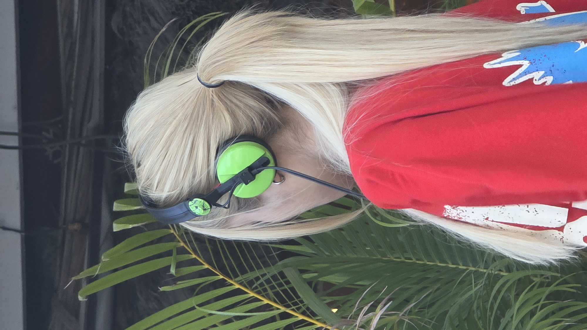 A person with long blonde hair pulled back in a ponytail is wearing green headphones and a red shirt with a blue and white design. They are standing amongst palm fronds. The location is unknown, and the specific city of Nha Trang, Vietnam is not discernable from this image.