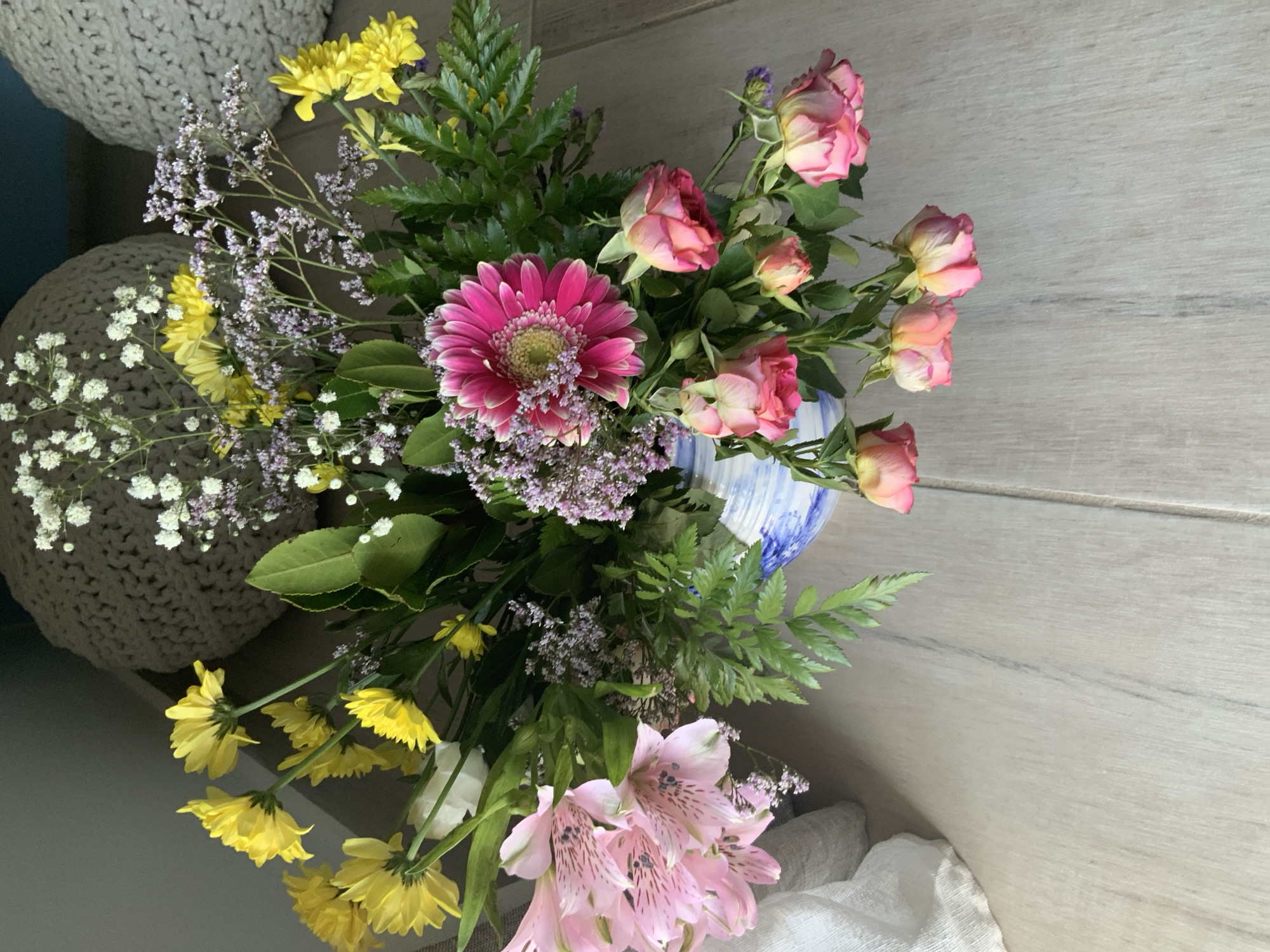 A bouquet of flowers, including pink alstroemeria, yellow chrysanthemums, a pink gerbera daisy, and small pink roses, is arranged in a blue and white striped vase. The vase sits on a light-colored wooden floor. A textured, grey pouf is visible in the background, as well as a white cloth. The scene appears to be indoors, likely in a domestic setting, possibly in Agios Nikolaos Municipal Unit, Greece, though this cannot be definitively confirmed from the image alone. The lighting suggests a daytime setting, and the overall impression is one of a naturalistic still life.