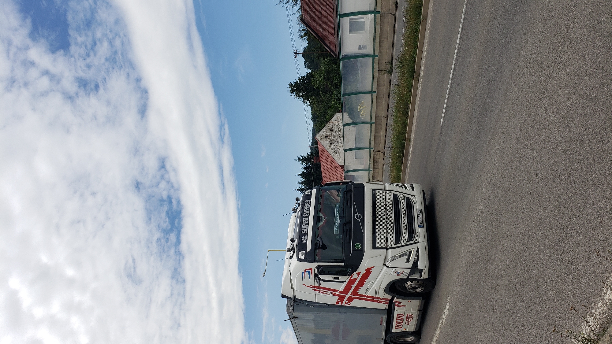 A white Volvo FH500 truck with "SUPER EXPRESS" branding and a red cross design on the side is driving on a road in Prešov, Slovakia. The truck has a grey trailer attached. In the background, there are houses with red roofs, trees, a concrete barrier with a green transparent shield, and a blue sky with white clouds. The truck's license plate is visible as "SU-PER02".