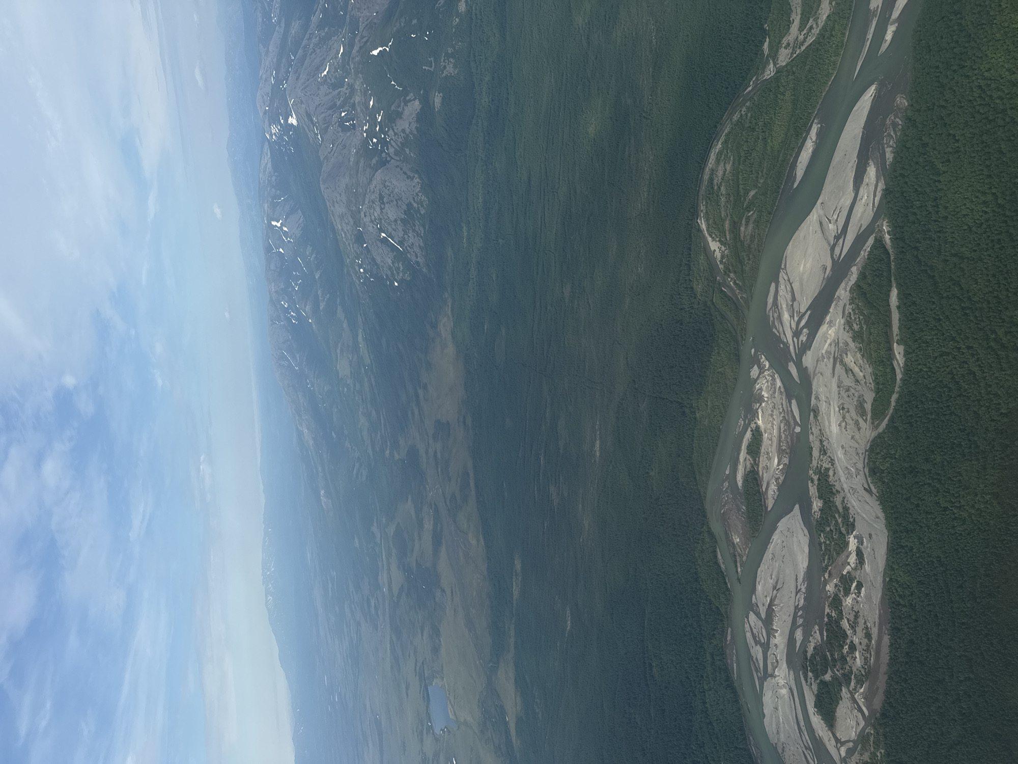 This aerial image depicts a vast, undeveloped landscape with a winding river system dominating the foreground. The river is braided, with numerous sandbars and islands covered in vegetation. Dense, dark green forests cover the banks of the river and extend into the middle ground. In the distance, rolling hills and mountains can be seen, some with patches of snow on their peaks. The sky is a pale blue with scattered white clouds. The image appears to be a natural, remote area, possibly in a region like Siberia or the Russian Far East, given the mention of Splavnaya, Russia, a city known for its river port and industrial activity. However, the depicted landscape does not show any urban development or human structures that would be indicative of Splavnaya itself, suggesting it is a view of the natural environment surrounding such a city rather than the city proper.
