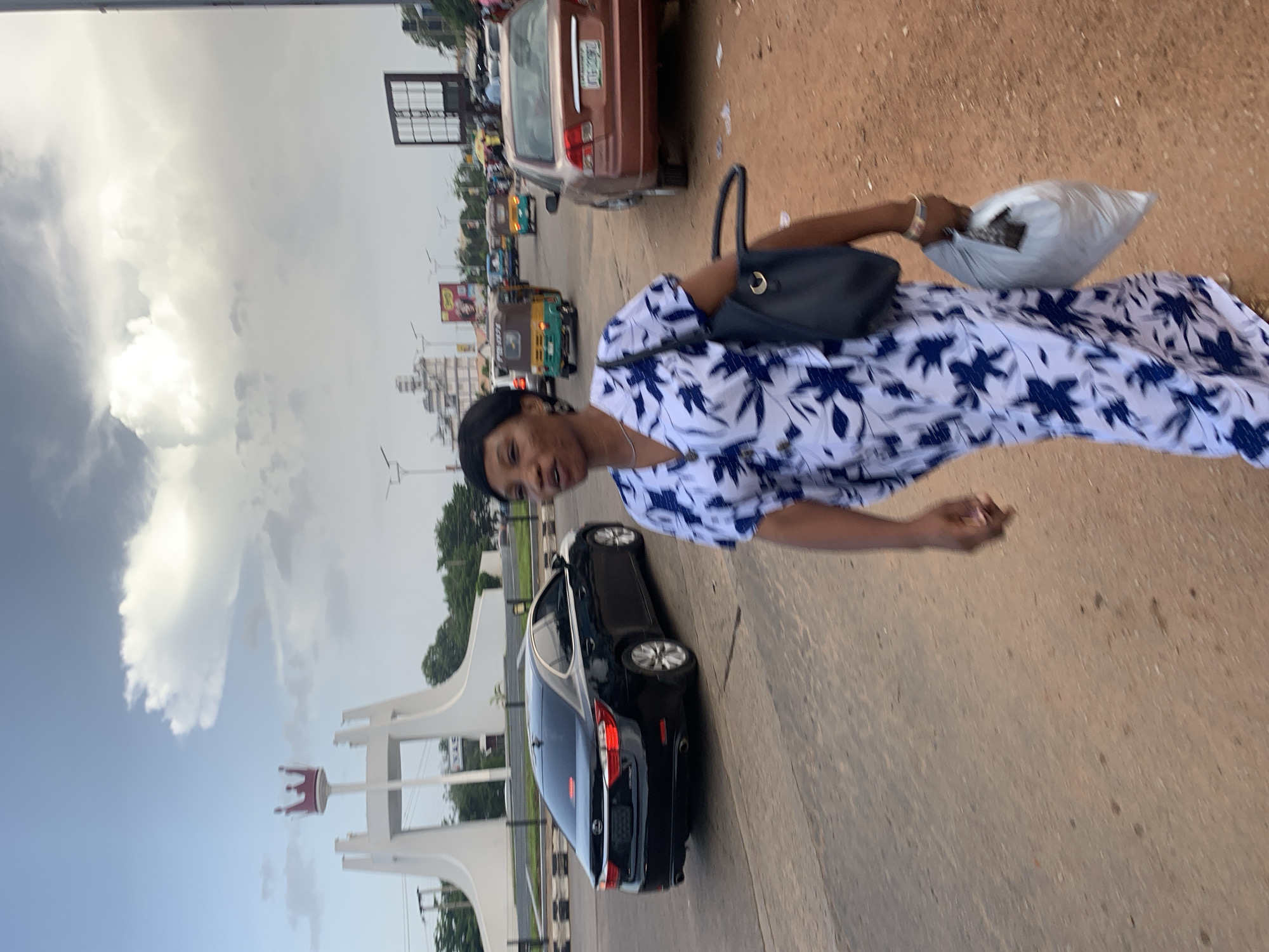 In the city of Jos, Nigeria, a woman walks on a road in front of a prominent monument. The monument is white and features a crown on top. The woman is wearing a white dress with a blue floral pattern and is carrying a black handbag and a white plastic bag. In the background, several vehicles, including cars and a tuk-tuk, are visible on the road. The sky is cloudy with patches of blue. The overall scene depicts an urban street in Jos.