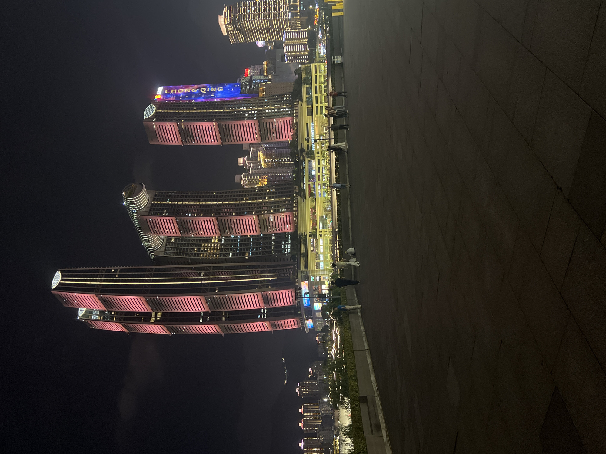 The image depicts a nighttime view of the Jiangbei Cheng Street (江北城街道) district in China, showcasing modern skyscrapers illuminated with pink and blue lights. In the foreground, a dark paved area, possibly a plaza or promenade, is visible with several individuals walking and standing. The middle ground features a multi-level structure with illuminated windows, gardens, and signs. Further in the background, a cityscape of numerous buildings with lit windows stretches across the horizon, with a body of water separating the foreground from the city. The sky is dark and overcast, typical of an urban night scene. The primary focus is on the architectural lighting of the prominent skyscrapers.