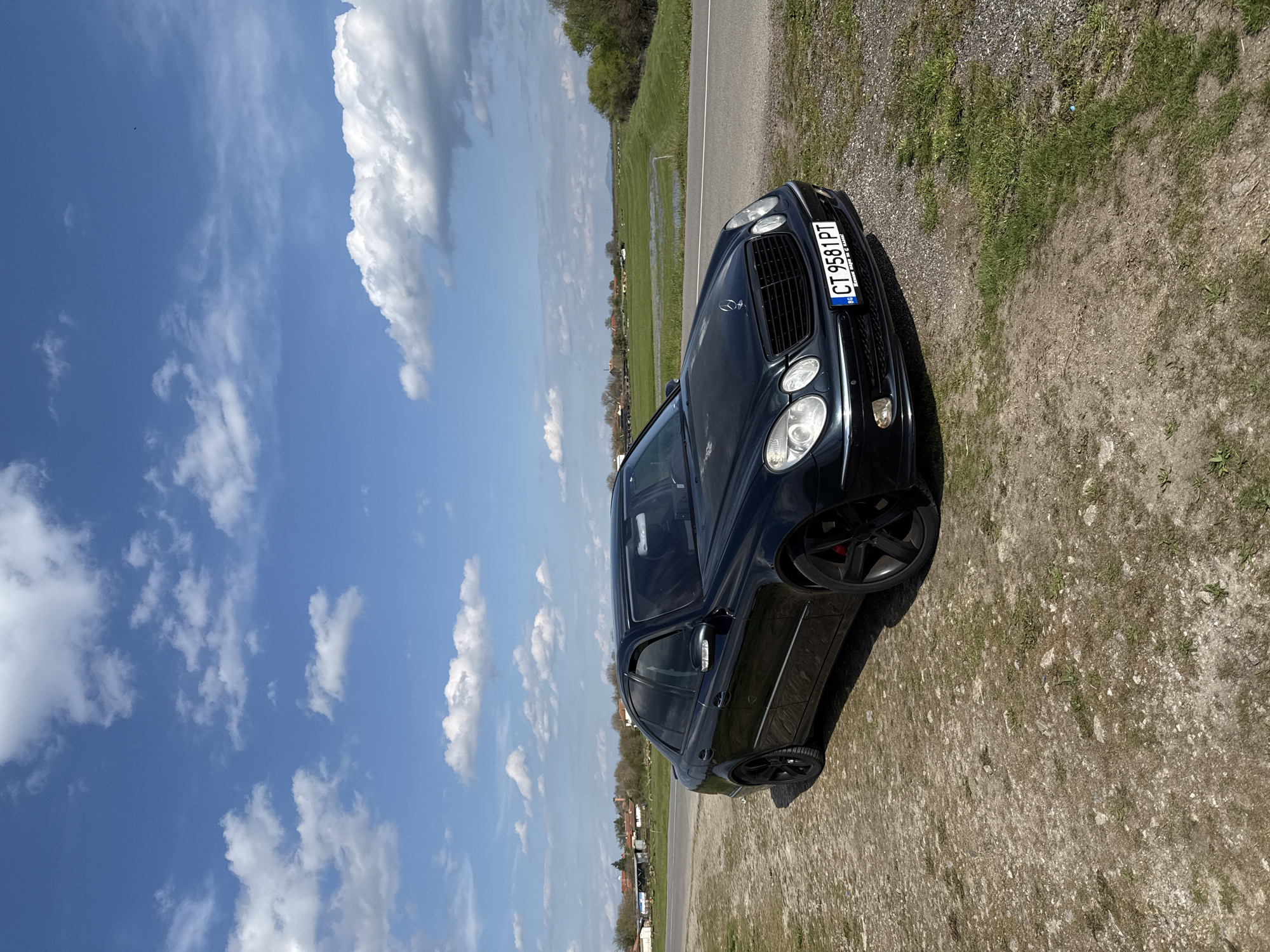 A black Mercedes-Benz E-Class, possibly a wagon, is parked on a dirt shoulder next to a paved road. The car is angled towards the viewer. The license plate reads "CT 9581 PT," suggesting the car is registered in Bulgaria. The car is parked near Pshenichevo, Bulgaria. A field with standing water and some vegetation stretches into the background. The sky is blue with scattered white clouds. Some small buildings or houses can be seen in the distant background. The ground near the car is a mix of dirt and patchy grass. The image is taken during daylight hours.
