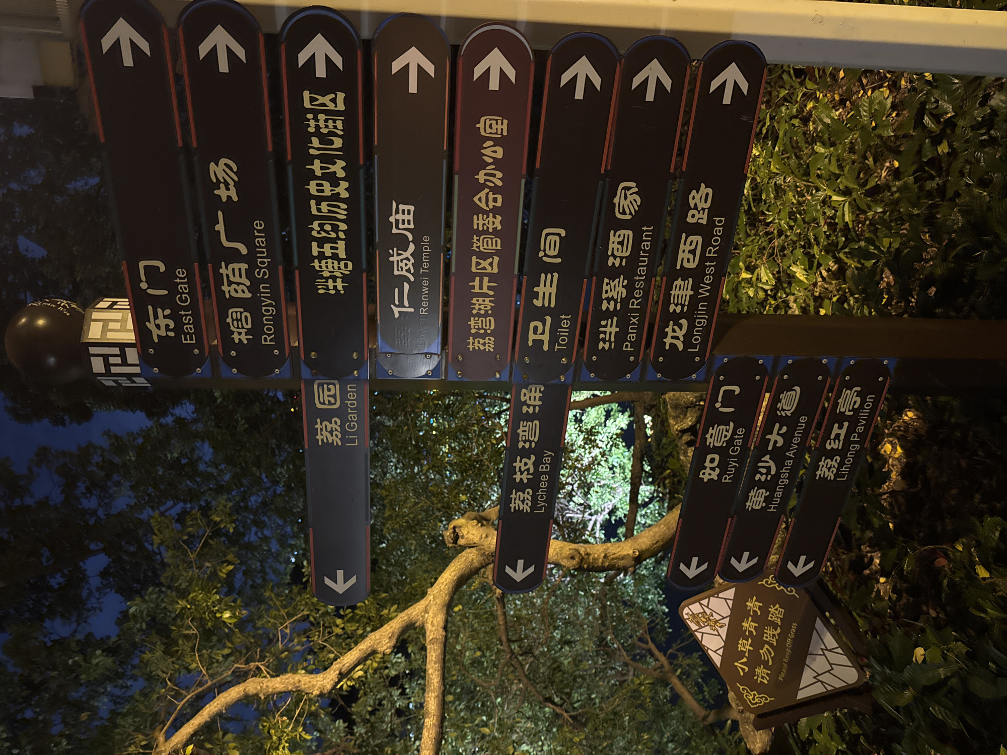 A photograph depicts a multi-directional street signpost in Liwan District, China. The signpost, affixed to a central pole, features multiple dark-colored rectangular signs with white directional arrows and text in both Chinese and English. The signs point towards various locations, including East Gate, Rongyin Square, Liwan History and Culture District, Li Garden, Renwei Temple, Lychee Bay, Liwan Lake Area Management Committee Office, Toilet, Panxi Restaurant, Longjin West Road, Ruyi Gate, Huangsha Avenue, and Lihong Pavilion. A smaller, diamond-shaped sign at the base reads "Please Keep Off Grass" in both Chinese and English. The surrounding environment includes lush greenery, trees, and hints of a dark evening sky.