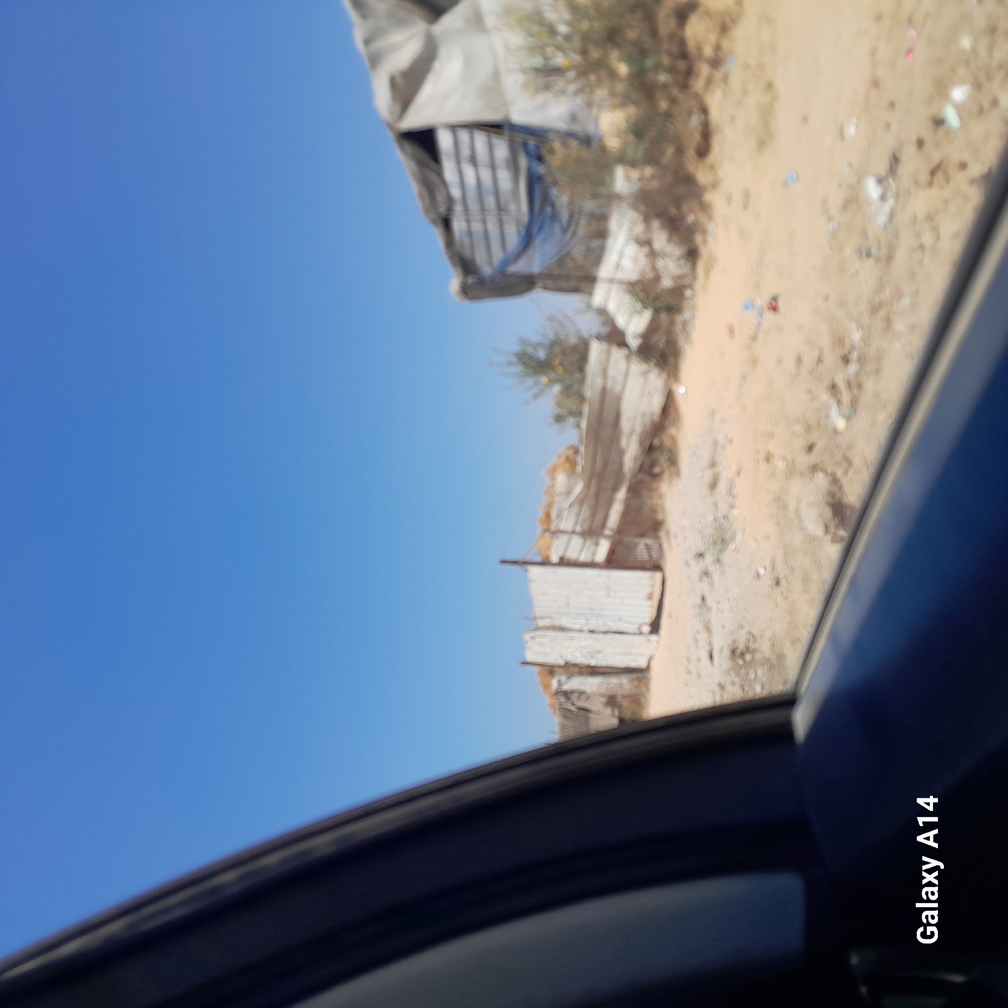 The image shows a view from inside a vehicle, looking out at a desolate, sandy area with several makeshift structures. The structures appear to be constructed from salvaged materials such as corrugated metal sheets, tarps, and wood. One structure on the right seems to be a large, partially collapsed shelter covered in a gray tarp, with visible metal framing underneath. To the left, there is a smaller structure with a corrugated metal wall and a gate. The ground is covered in sand, with scattered debris including plastic bottles and other trash. Sparse, dry vegetation can be seen in the background. The sky is a clear, bright blue. The scene suggests an informal settlement or a place of temporary shelter in an arid environment, consistent with the description of Deir el-Balah, Palestinian Territories. The watermarks in the image indicate it was taken with a "Galaxy A14".