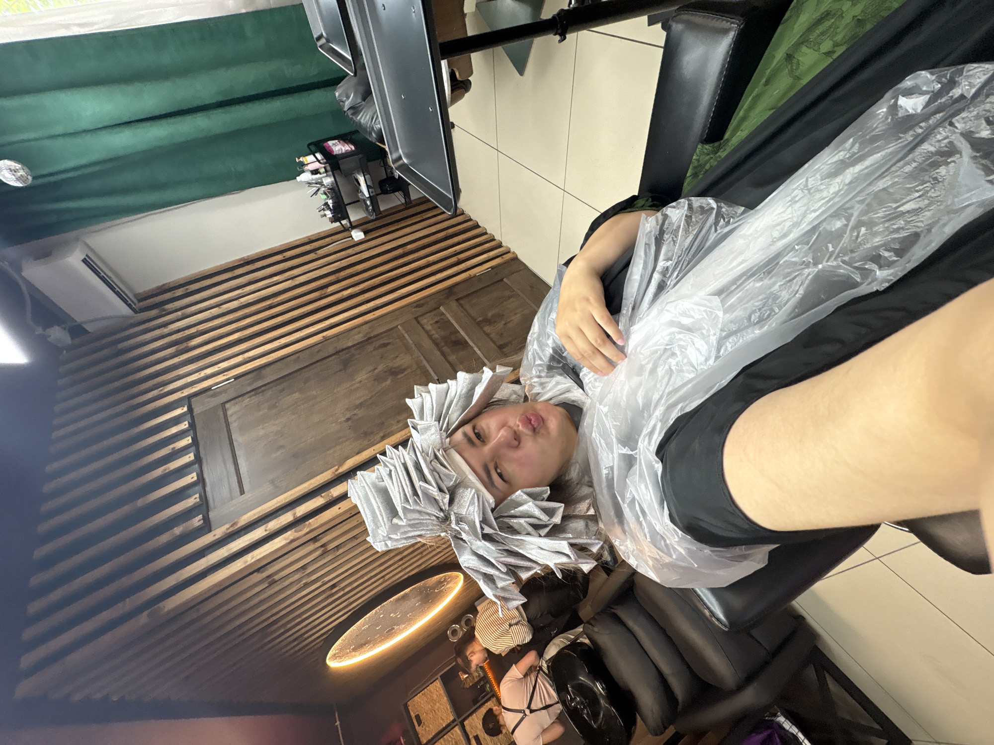 A person is seated in a salon chair, their head wrapped in numerous foil packets, likely for hair treatment. They are wearing a plastic cape to protect their clothing. The person is looking at the camera and puckering their lips. In the background, other salon patrons are visible, and the salon features wooden paneling on the walls and a disco ball hanging from the ceiling. The scene suggests a hair salon in Birobidzhan, Russia, as indicated by the provided image context.