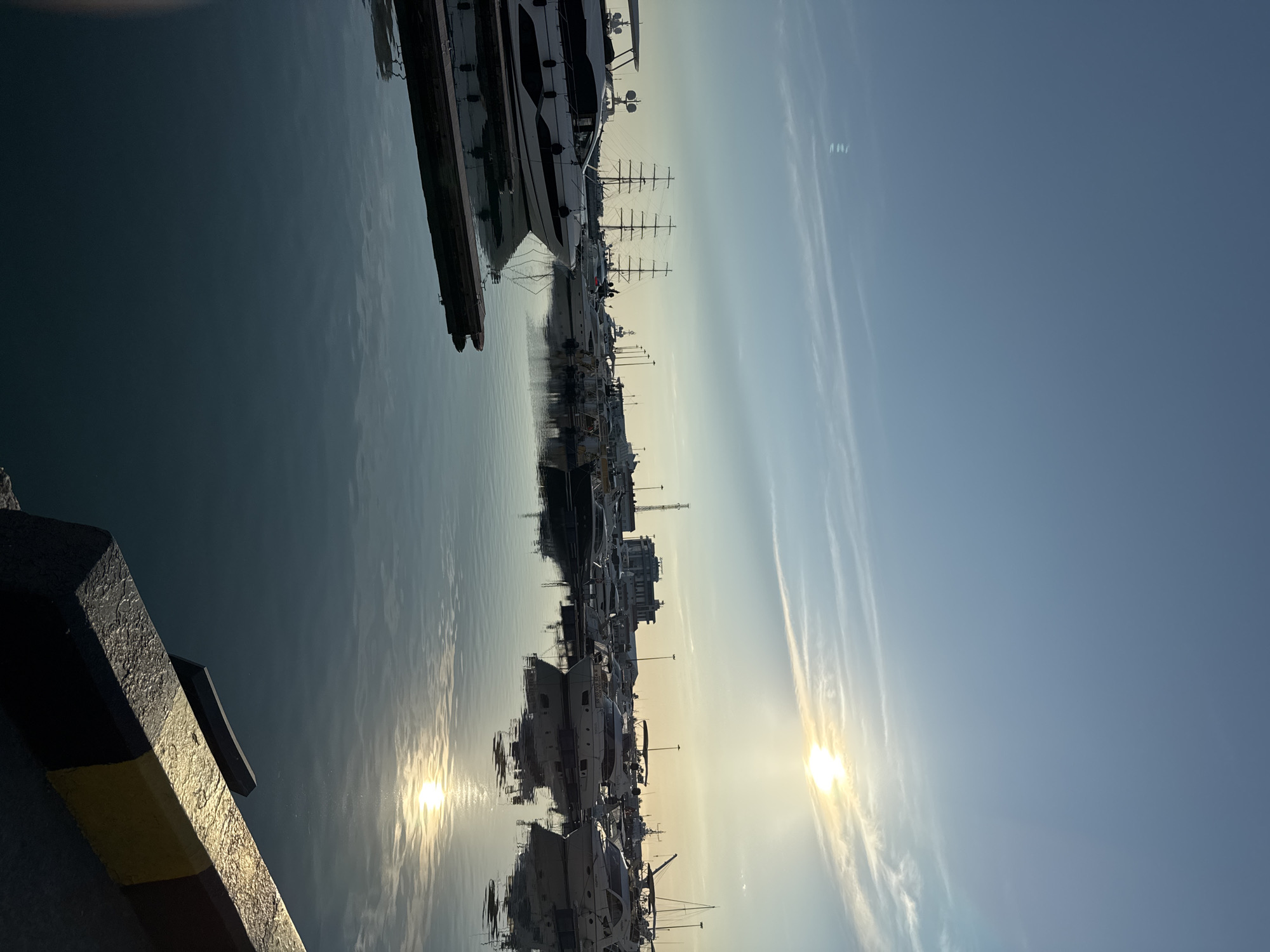 This is a vertical, medium-shot photo taken at a marina in Sochi, Russia. The scene depicts a row of yachts and boats docked in the water, with their reflections visible on the calm surface. The sun is low in the sky, casting a warm, golden light and creating a bright glare on the water. The sky is a clear blue with wispy white clouds, suggesting it is either early morning or late afternoon.
The foreground of the image shows the edge of a dock with yellow and black markings. Beyond the dock, several large yachts are lined up. The most prominent is a large, white yacht with dark accents, featuring multiple decks and a sleek design. Behind it, a sailing yacht with tall masts is visible. Further down the line, more boats of various sizes are moored.
The overall mood of the image is peaceful and serene, enhanced by the soft lighting and the stillness of the water. The sun's glow suggests a pleasant day for being out on the water. There is no visible text in the image.