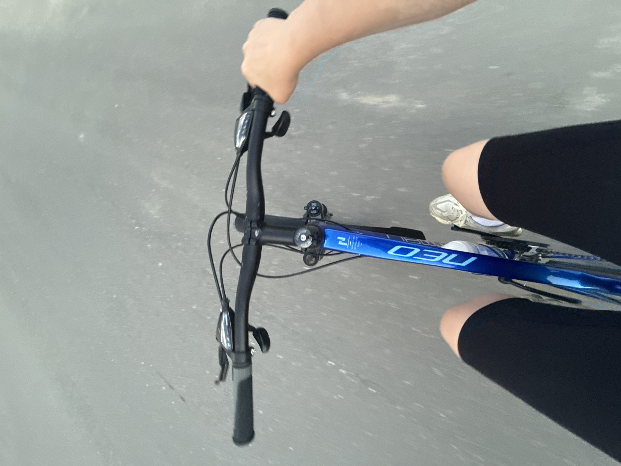 A first-person, downward-angle photograph captures an individual riding a blue bicycle on a paved surface. The image exhibits significant vertical motion blur in the background, indicating forward movement.
In the foreground, black bicycle handlebars are visible, fitted with black grips, silver and black brake levers, and gear shifters. A person's right hand grasps the right handlebar. The bicycle's frame is a metallic blue, with the white text "NEO" inscribed vertically on the down tube. A white water bottle is secured within a cage on the bicycle frame.
The rider's upper thighs and knees are visible on both sides of the bicycle frame, clad in black shorts or cycling leggings. The right foot, wearing a white and grey athletic shoe, is partially visible near the pedal area. The paved surface beneath the bicycle appears grey with streaks from motion blur. The scene is located in the city of Murom, Russia.