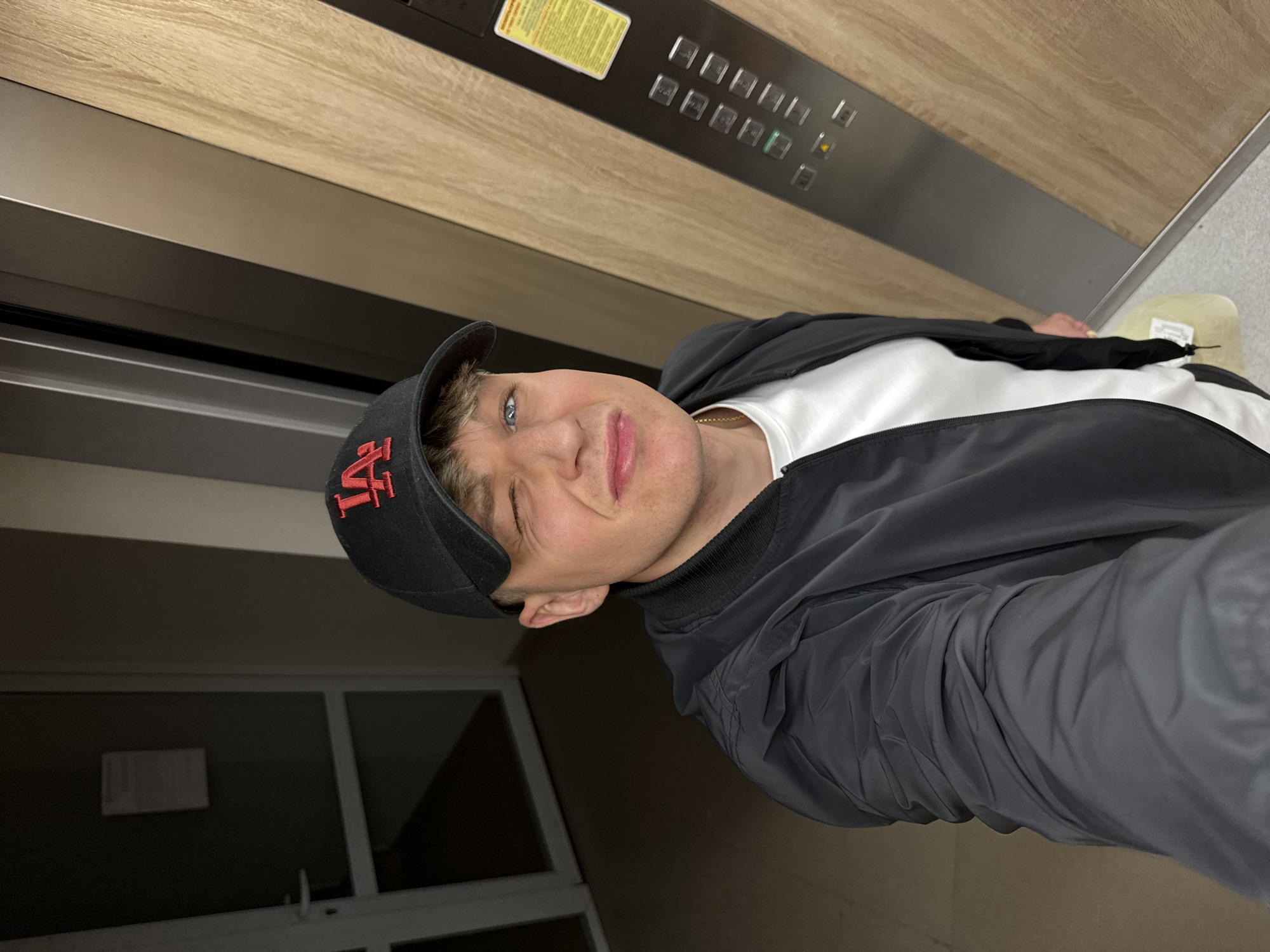 A young adult male, appearing in a selfie, is positioned inside a modern building in Plovdiv, Bulgaria. He has light brown hair, blue eyes, and is winking with his left eye while exhibiting a slight smirk. He wears a black baseball cap featuring a red "LA" logo, a black zippered bomber jacket over a white t-shirt, and a thin gold chain around his neck. To the subject's left, a white-framed door with a dark panel and a white handle is visible in a dimly lit hallway, with a rectangular paper note affixed to its upper portion. To the subject's right, a contemporary elevator interior or adjacent wall is composed of vertical dark grey/metallic and light wood-grain panels. A silver elevator control panel on the right features multiple square buttons, with one button illuminated green and another amber, alongside a yellow rectangular warning or information sticker near the top. The floor is light grey, and a pale, possibly yellowish, object rests on it near the subject's lower right.