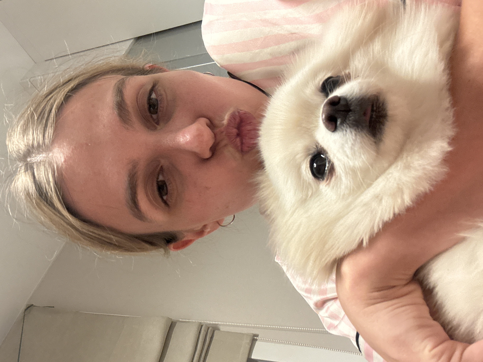 An indoor close-up image depicts an adult female holding a small white dog. The female, positioned in the upper portion of the frame, has light-colored hair, fair skin, and is wearing a pink and white striped garment with black trim. She has small hoop earrings and her lips are pursed in a kissing gesture, directed towards the camera. Her right arm is visible, supporting the dog. The dog, a fluffy white breed with dark eyes and nose, is held in the lower center of the frame, close to the female's chest. A small portion of its pink tongue is visible. The background reveals a light-colored wall and ceiling, with a window fitted with beige horizontal blinds and a vertical beaded pull chain visible on the left side. The lighting is even, suggesting an interior residential setting in Unknown, Unknown.