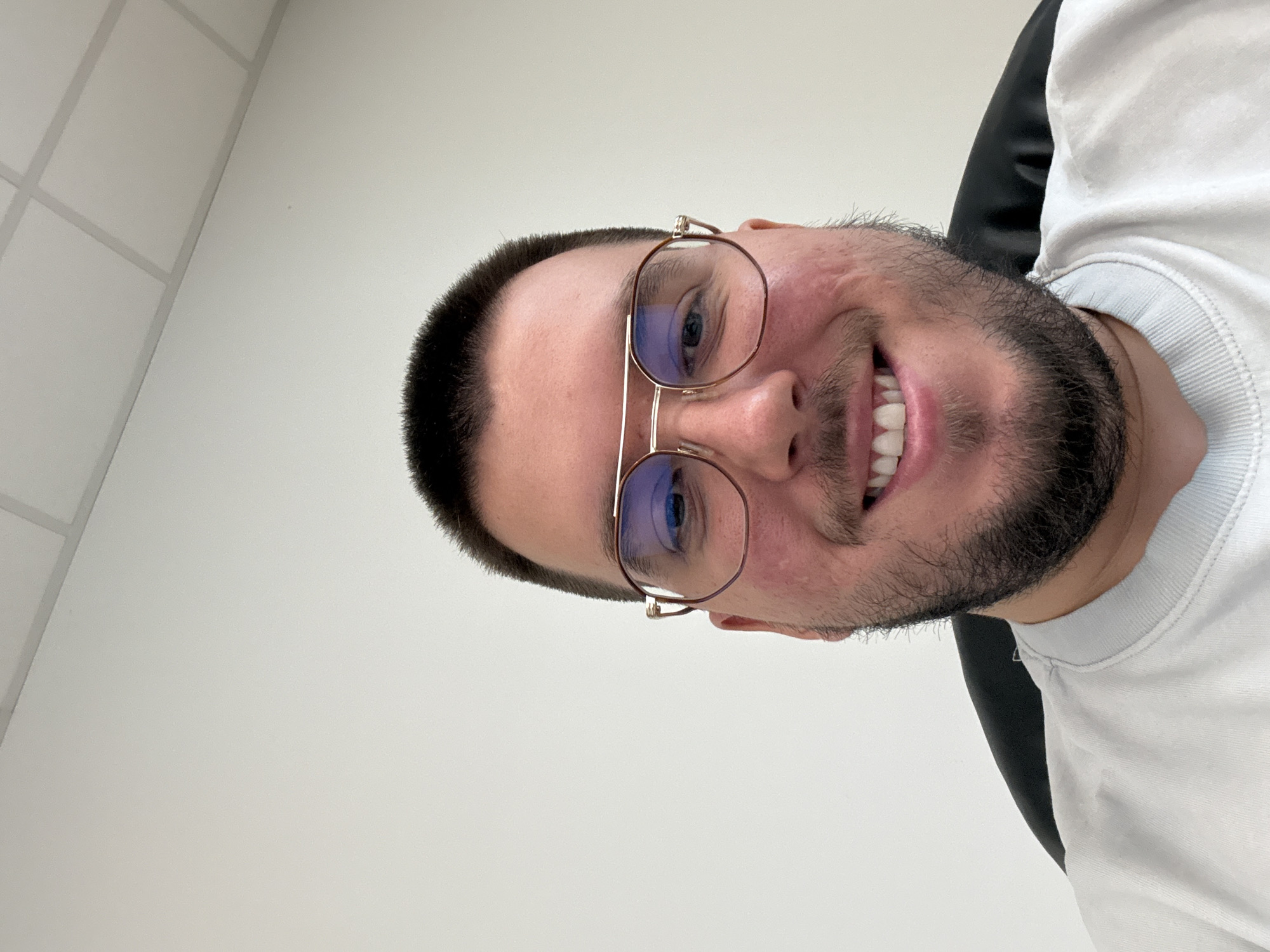 A man with short dark hair and a beard is smiling at the camera while wearing gold-rimmed glasses and a white sweatshirt. He is sitting in a black chair. The background is a white wall, likely indoors. There is no information in the image that indicates it was taken in Sofia, Bulgaria.