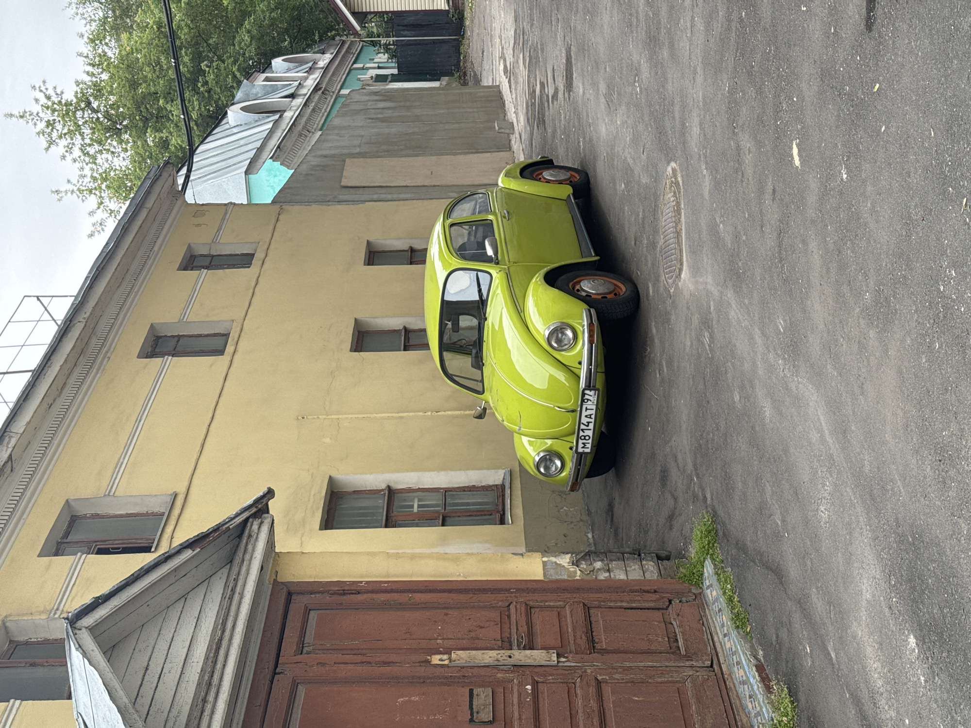 The image shows a bright green Volkswagen Beetle parked on a narrow, paved alleyway in Moscow, Russia. The car is the primary object of focus, positioned diagonally across the frame. The building to the left is a pale yellow, multi-story structure with several windows. To the right of the car is a gray asphalt surface with a manhole cover. Overhanging trees with green foliage are visible at the top of the image, suggesting it is daytime and likely spring or summer. The overall scene appears to be an urban environment, possibly a courtyard or a back street, characterized by older architecture. There is a wooden door in the foreground with a weathered appearance. Visible text on the car's license plate reads "M814AT 97". The lighting suggests it is daytime, and the sky appears overcast.