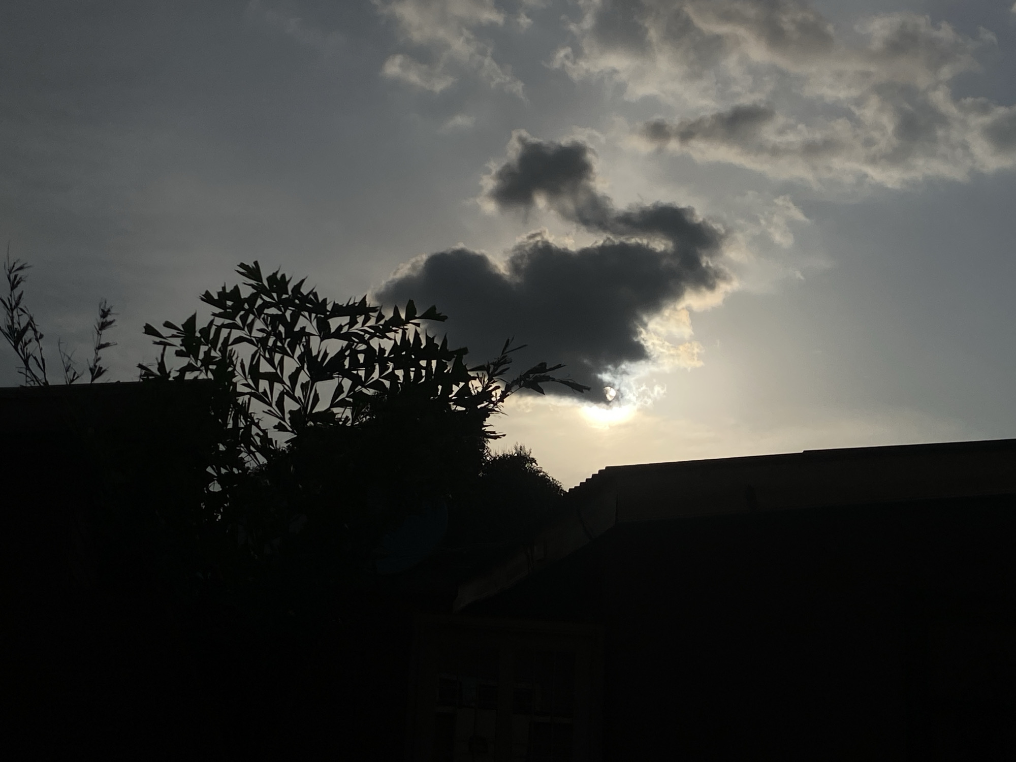 The image depicts a twilight scene with silhouettes of trees and rooftops against a cloudy sky. The sun is partially obscured by dark clouds, casting a soft glow. The foreground features dark, silhouetted foliage and the dark shapes of buildings, possibly residential structures, with a hint of windows visible on one. The overall atmosphere is serene and somber. No people are visible in the image. The location is Barden Yana, Nigeria.