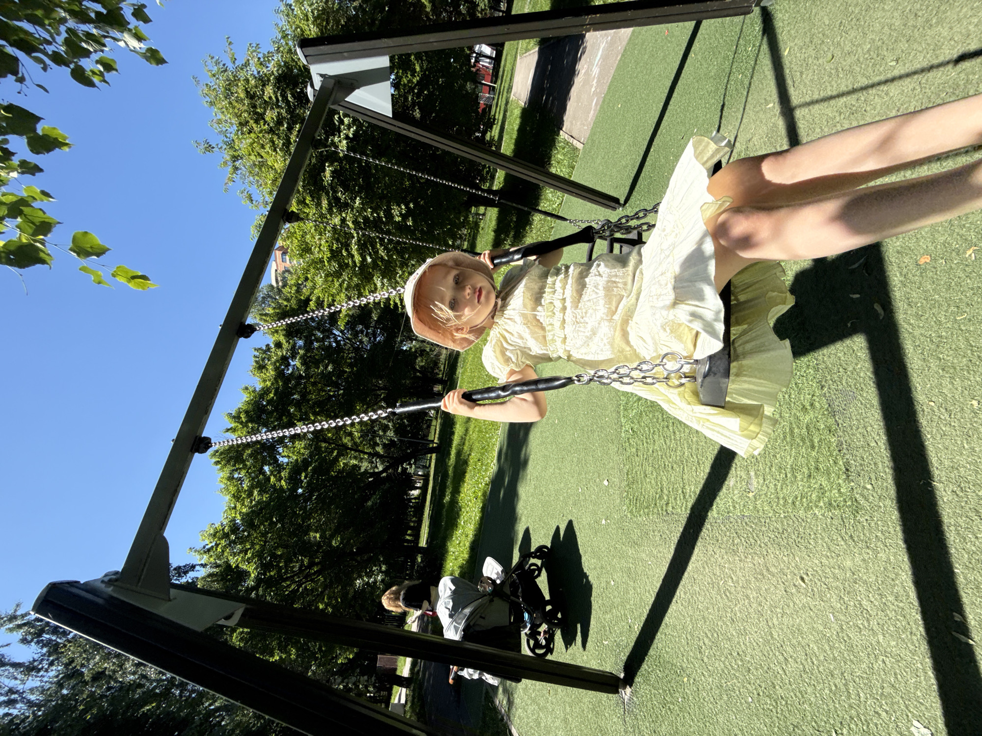 The image depicts a young blonde child on a swing in a park located in Moscow, Russia, under a clear blue sky. The child, dressed in a light yellow ruffle dress and a light pink wide-brimmed hat, is seated on a swing with black handles and metal chains, looking directly towards the camera. The swing set consists of dark-colored metal A-frame supports and a horizontal beam. The ground surface immediately surrounding the swing set is green synthetic turf, transitioning to natural grass further into the park. Numerous deciduous trees with vibrant green foliage fill the background. To the left of the main subject, an adult is partially visible next to a light grey baby stroller. A paved pathway can be seen winding through the park in the mid-ground. Bright sunlight creates distinct shadows from the swing structure and the child, indicative of a sunny day.