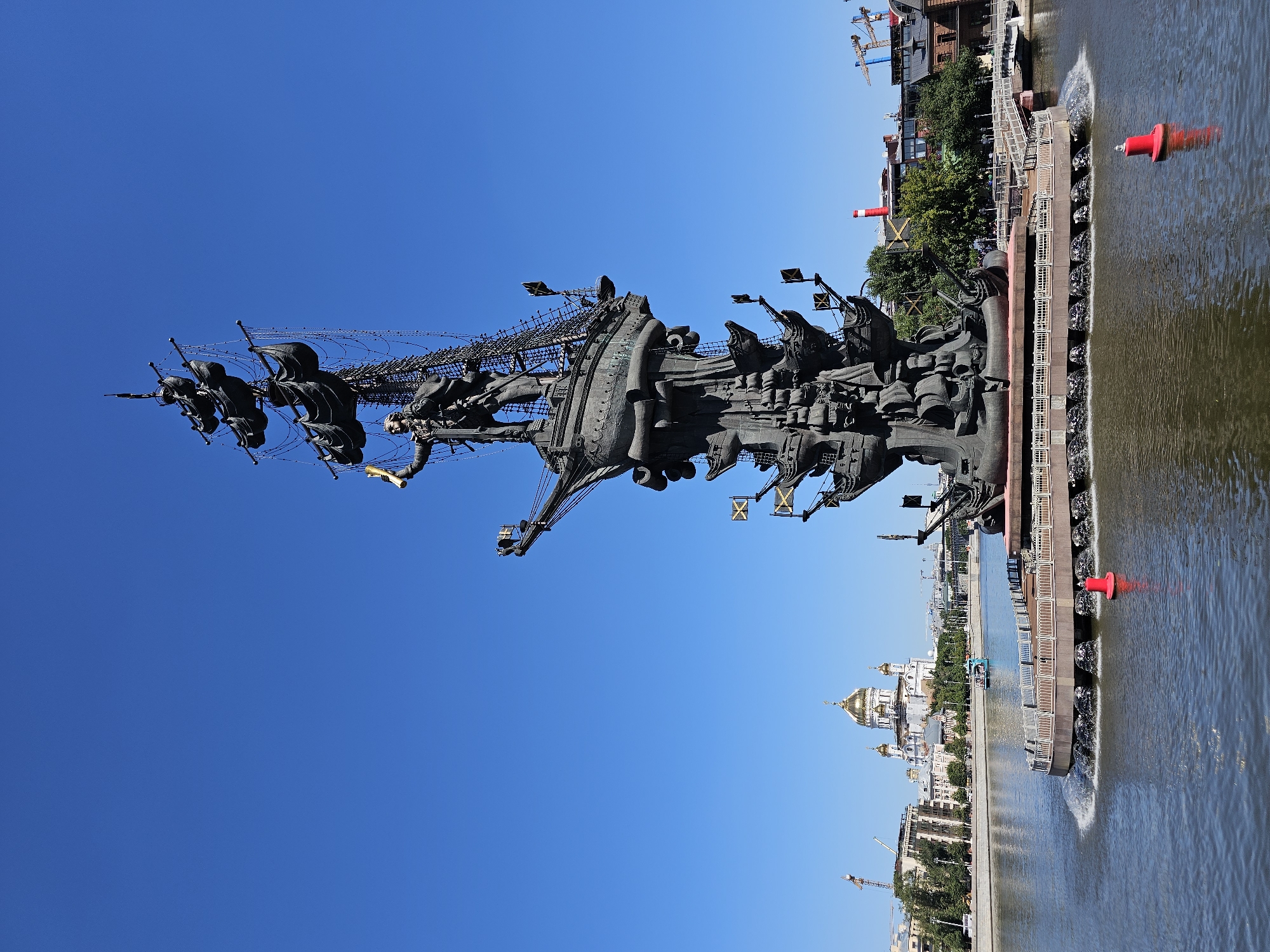 The image depicts the Peter the Great Statue in Moscow, Russia, situated on an artificial island in the Moskva River. The colossal bronze statue, designed by Zurab Tsereteli, stands tall, commemorating Peter the Great's naval reforms and his role in Russia's modernization. The statue is composed of several ship-like structures stacked upon one another, culminating in a figure of Peter the Great holding a golden scepter. Several smaller ships are depicted as part of the base, adorned with flags. The statue is surrounded by a fountain that creates a spray of water around its base. In the background, the Moscow skyline is visible, featuring the golden domes of the Cathedral of Christ the Saviour and other buildings. The sky is clear and blue, indicating a sunny day. The river reflects the blue sky and the surrounding structures. There are no people clearly visible in the frame, except for figures on a distant boat.