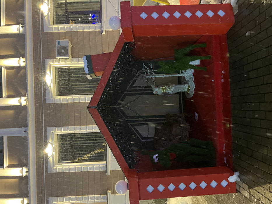 The image displays an illuminated outdoor Christmas decoration at night during a snowfall, located in Terjola, Georgia. The central subject is an open-front, red, house-like structure featuring a black gabled roof with red trim and a small red chimney topped with white and green elements on the right side. Red columns with white diamond insets frame the front, each capped with a white spherical light. The interior floor is red, and the rear wall exhibits vertical dark stripes, adorned with white string lights. Inside the display, from left to right, are a large, green, stylized figure with white eyes, an ornate brown armchair, a standing white and gold figure statue on a white base, a bare white artificial tree, and a smaller green artificial deer. In the background, a light-colored building with multiple windows framed by white decorative moldings is visible. Upward-facing lights illuminate the building's facade. An external air conditioning unit is mounted between two windows on the right. The foreground consists of dark paver bricks, with small accumulations of snow present around the base of the Christmas display. Snowflakes are actively falling and visible across the entire scene.