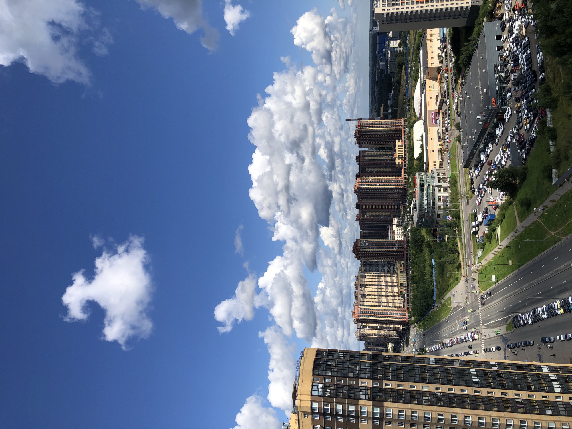 This is a high-angle, outdoor, daytime shot of a city scene. In the foreground, a tall, modern residential building with a beige facade and numerous windows dominates the left side of the frame. Across the street, a multi-lane road runs diagonally away from the viewer, lined with parked cars and a few pedestrians. Beyond the road, a row of similar tall, modern residential buildings, predominantly orange and beige, stretches across the mid-ground. Further back, various commercial buildings and green spaces are visible, including a large building with a sign that reads "LADA." In the background, a flat, developed cityscape extends to the horizon. The sky is a vibrant blue, populated by numerous white, fluffy cumulus clouds.