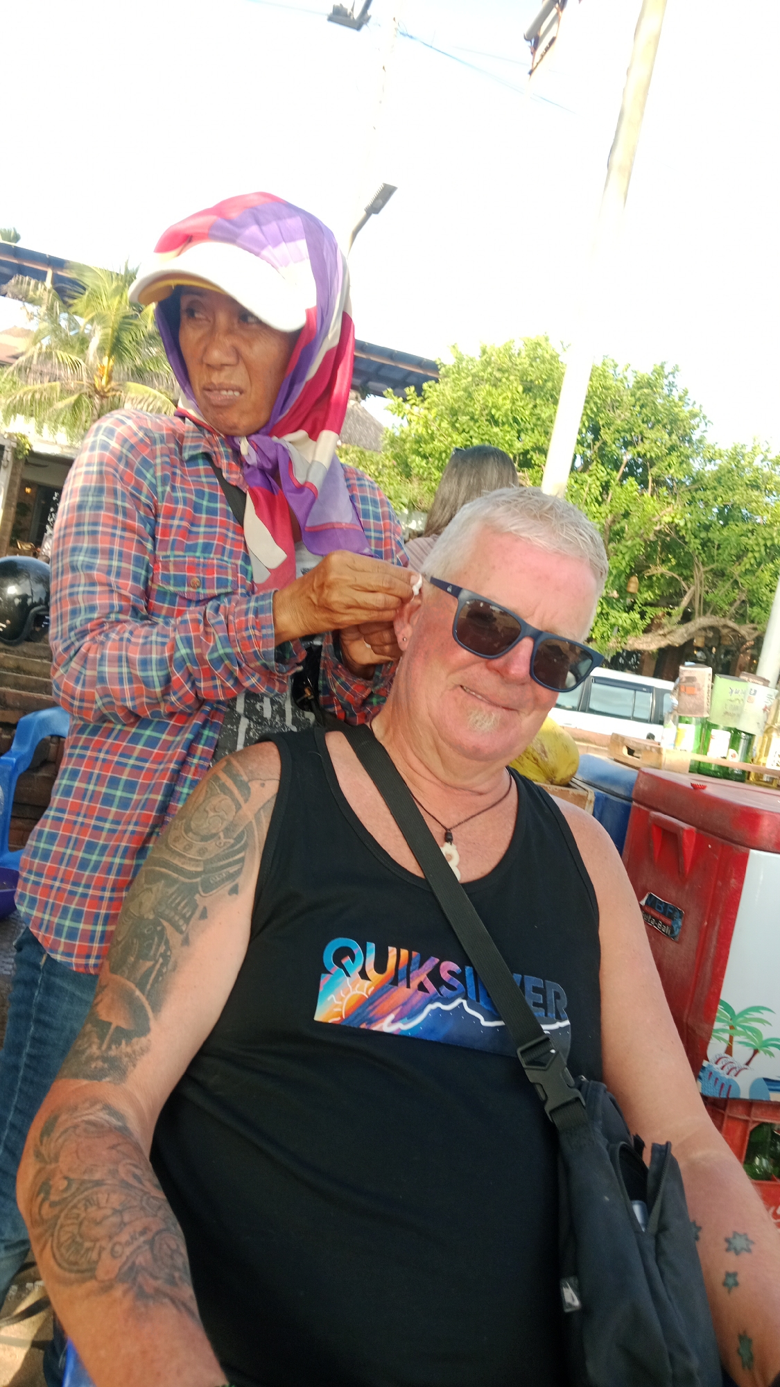 In Kuta, Indonesia, an individual with grey hair and sunglasses is having their ear cleaned by another person. The person cleaning the ear is wearing a plaid shirt and a headscarf. The man in sunglasses is wearing a black tank top with a Quiksilver logo and has tattoos on his arm. The background shows lush green trees and a bright sky, suggesting an outdoor setting.