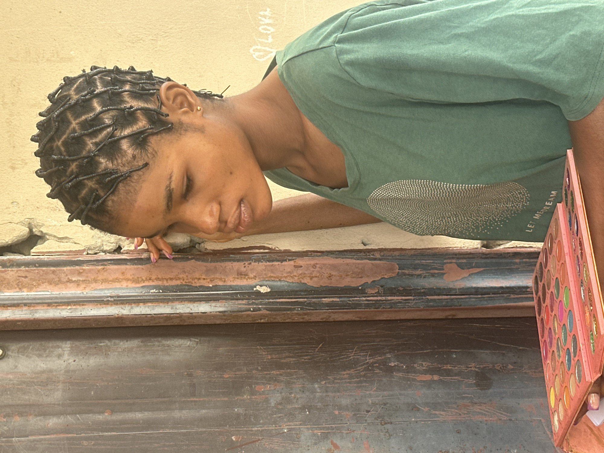 A young Black woman with braided hair is leaning against a weathered, dark brown wooden door. She is looking down with her right hand touching her cheek. She wears a green t-shirt with a silver circular design and some text. In the foreground, a large pink makeup palette with many eyeshadows is visible. The background consists of a textured, light beige wall with some white chalk writing that appears to spell "LOVE" with a heart. The scene is likely located in Badagry, Nigeria, given the architecture and the presence of the woman.