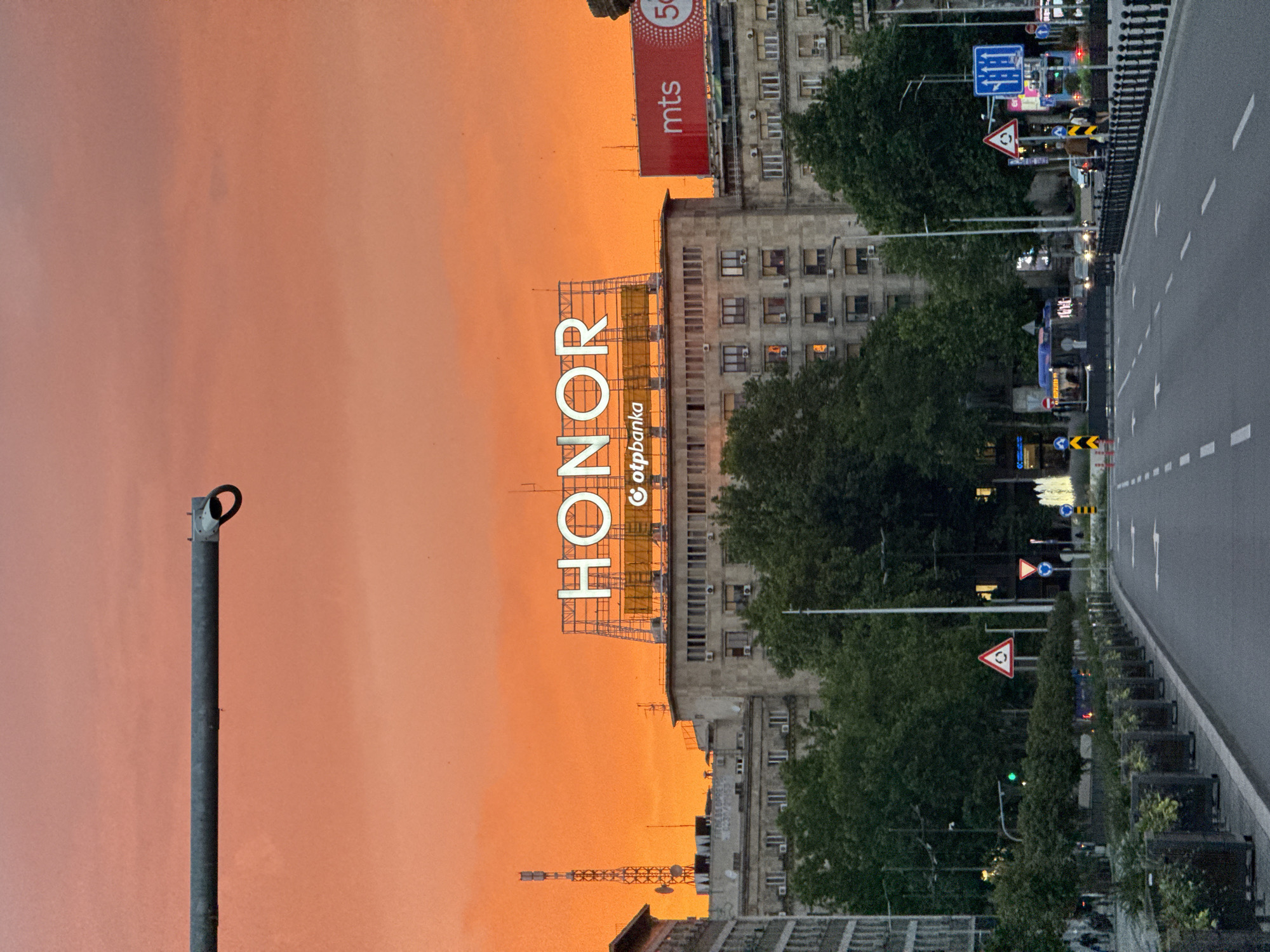 This image captures a city scene in the Vracar Urban Municipality, Serbia, during what appears to be sunset or sunrise, given the vibrant orange sky. In the foreground, a paved road with white lane markings leads into the distance. Along the left side of the road, planters with greenery line the sidewalk, and a light pole extends from the top left of the frame.
In the mid-ground, a large, multi-story building dominates the view, with numerous windows visible. A prominent neon sign displaying "HONOR" in large white letters is mounted on top of this building, with a smaller sign for "otpbanka" underneath. To the right of the "HONOR" sign, another building features a large red billboard with the white text "mts" and a circular logo. Trees with dense green foliage are situated in front of the buildings.
Further back, a tall telecommunications tower is visible to the left. The street is populated with a few cars and a bus, and several traffic signs are placed along the roadside. People are present on the sidewalks and crossing the street. The overall atmosphere is that of a busy urban environment under a striking sky.