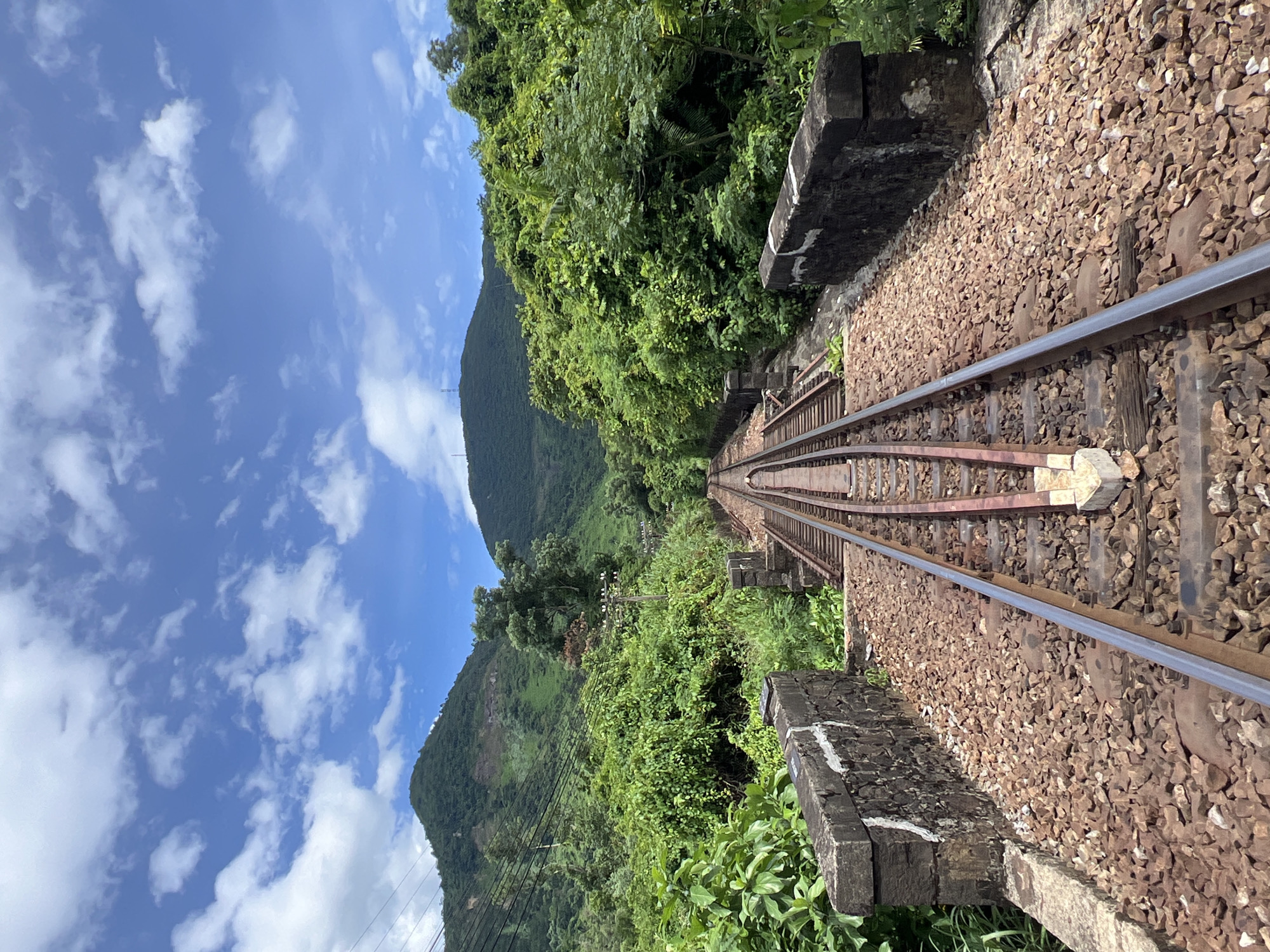 The image shows a set of railroad tracks receding into the distance, flanked by lush green vegetation and rolling hills under a bright blue sky with scattered white clouds. The tracks appear to be part of an old railway bridge with stone abutments on either side, visible in the foreground. The scene is set in a natural, somewhat overgrown environment, suggesting a rural or semi-rural location. Given the context of Huế, Vietnam, this could be a section of the Reunification Express line, which passes through scenic landscapes on its route, potentially near the Hai Van Pass or other coastal mountain ranges surrounding the city. There are no people or animals visible in the image, and no vehicles are present on the tracks. The lighting indicates it is daytime, likely mid-morning or afternoon. The overall impression is one of a tranquil, somewhat secluded railway line amidst vibrant natural scenery.