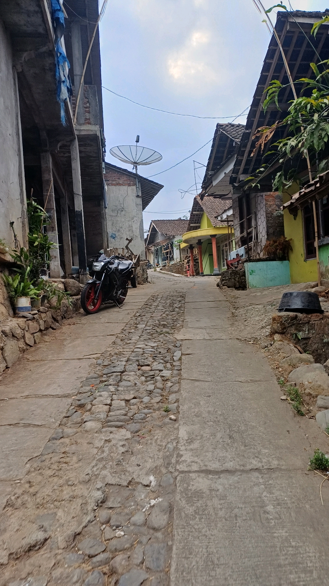 The image depicts a narrow, upward-sloping street in Wonokerso, Indonesia, characterized by a mix of cobblestone and concrete paving. The scene is lined with houses of varying designs and colors, some appearing unfinished or in disrepair, others displaying brighter hues like yellow and green. Vegetation, including potted plants and larger trees, is visible along the sides of the street. A black motorcycle with red accents is parked on the left side of the road. A satellite dish is mounted on the roof of one of the buildings. The sky is overcast, suggesting either early morning or late afternoon. The overall impression is one of a rural or semi-urban residential area.