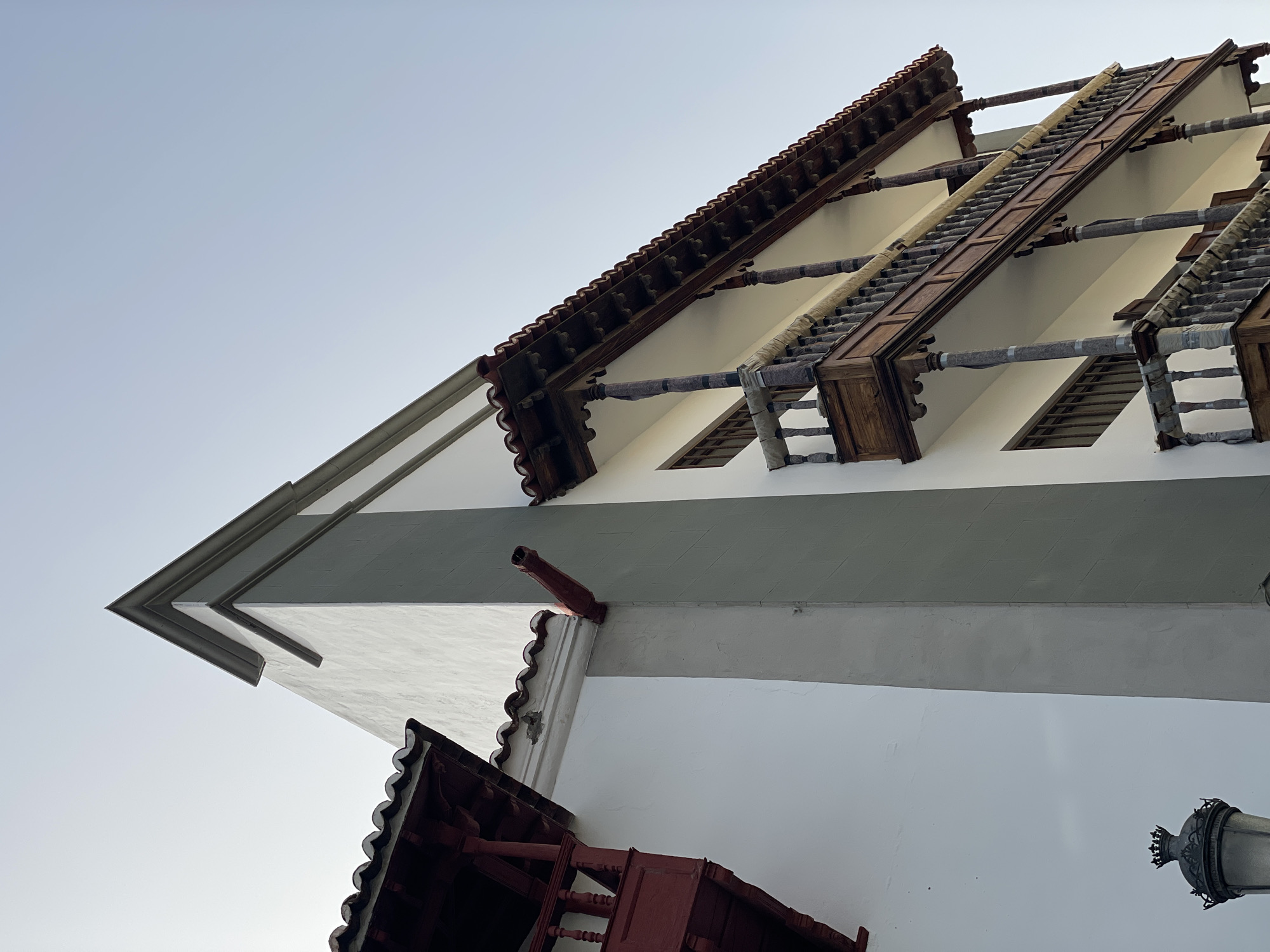 The image shows a low-angle view of a building facade in Santa Cruz de Tenerife, Spain. The architecture features white walls with a broad gray horizontal band. There are visible sections of the building's roof, which consist of reddish-brown tiles and exposed dark wooden beams and supports. Some of the wooden structures appear to be wrapped in a light-colored padding, potentially for protection. The sky is pale blue and mostly clear. At the bottom right of the image, a portion of a vintage-style streetlight is visible. A red gutter and roof overhang are also present.