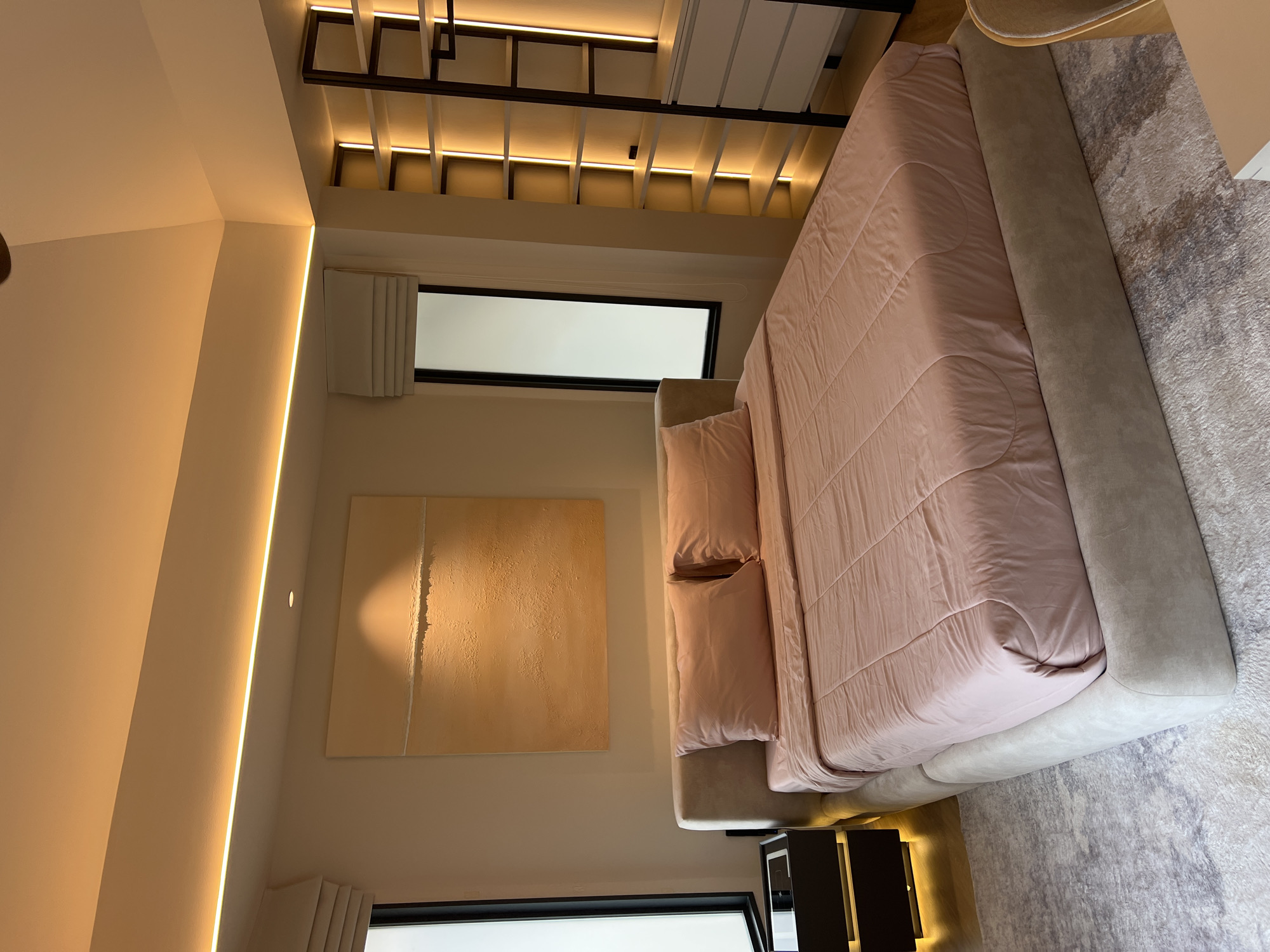The image shows a bedroom interior. The bed is covered with a light pink duvet and matching pillows, and has a gray upholstered headboard. A light gray carpet is partially visible under the bed. Two dark-colored bedside tables are next to the bed. Above the bed, a window is visible. On the wall, there is an abstract artwork in beige and white tones. On the ceiling, there is a decorative wooden structure with integrated lighting. A vertical strip of light runs along the wall on the left side. The location appears to be Baan Bang Rak, Thailand, based on the overall architectural and interior design style.
