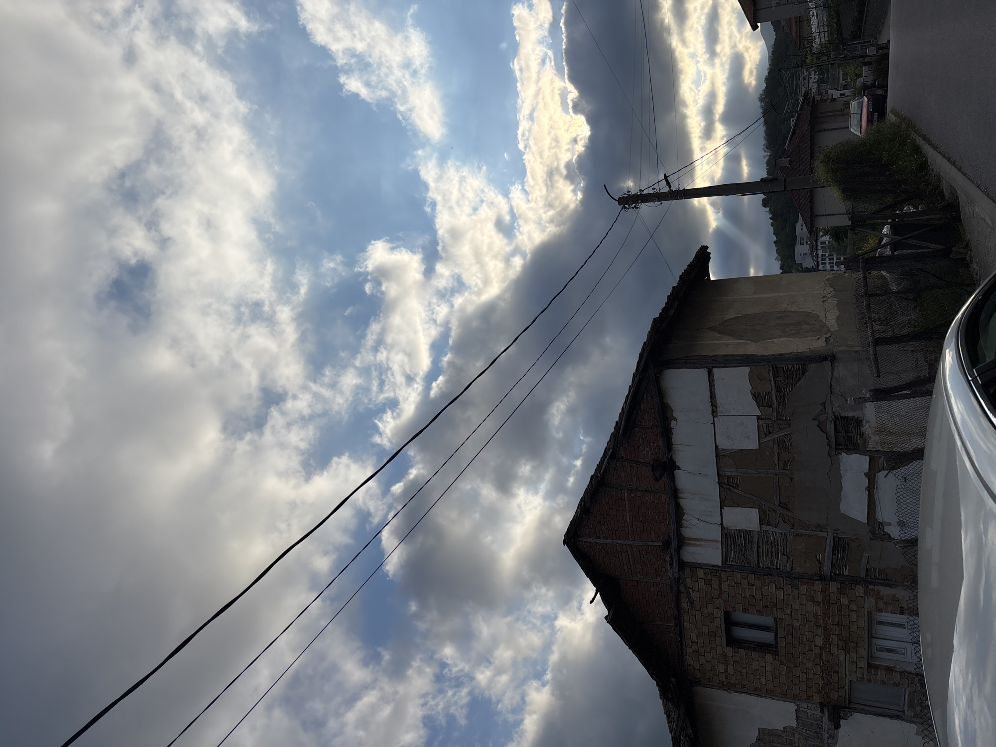 The image shows an upward-angled street view in Zlatograd, Bulgaria. A dilapidated brick and plaster building is featured on the right, appearing worn and partially decayed. Power lines extend across the upper portion of the image. The sky contains a mix of blue patches and dense, layered clouds, partially illuminated by sunlight. A section of a white vehicle is visible in the bottom right corner. A telephone pole is visible. Additional buildings can be seen in the distance.