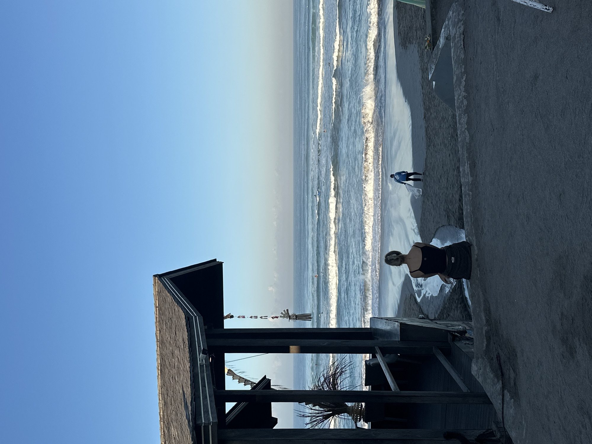 In North Kuta, Indonesia, an outdoor scene captures a serene beach setting under a clear blue sky. In the foreground, a covered structure with a shingled roof and dark wooden posts frames the view of the ocean. To the left of the structure, a tall, thin plant in a woven planter adds a touch of greenery.
In the mid-ground, a woman is seated on a concrete ledge, facing the ocean. She has blonde hair pulled back and wears a dark tank top and dark pants. A small stream of water flows from behind her towards the beach.
Further out, on the sandy shore, a man walks towards the water, carrying a white surfboard. He is dressed in a blue long-sleeved shirt and dark pants. The ocean exhibits gentle waves, and several surfers are visible scattered across the water. In the distance, the horizon is clear, meeting the expansive blue sky.