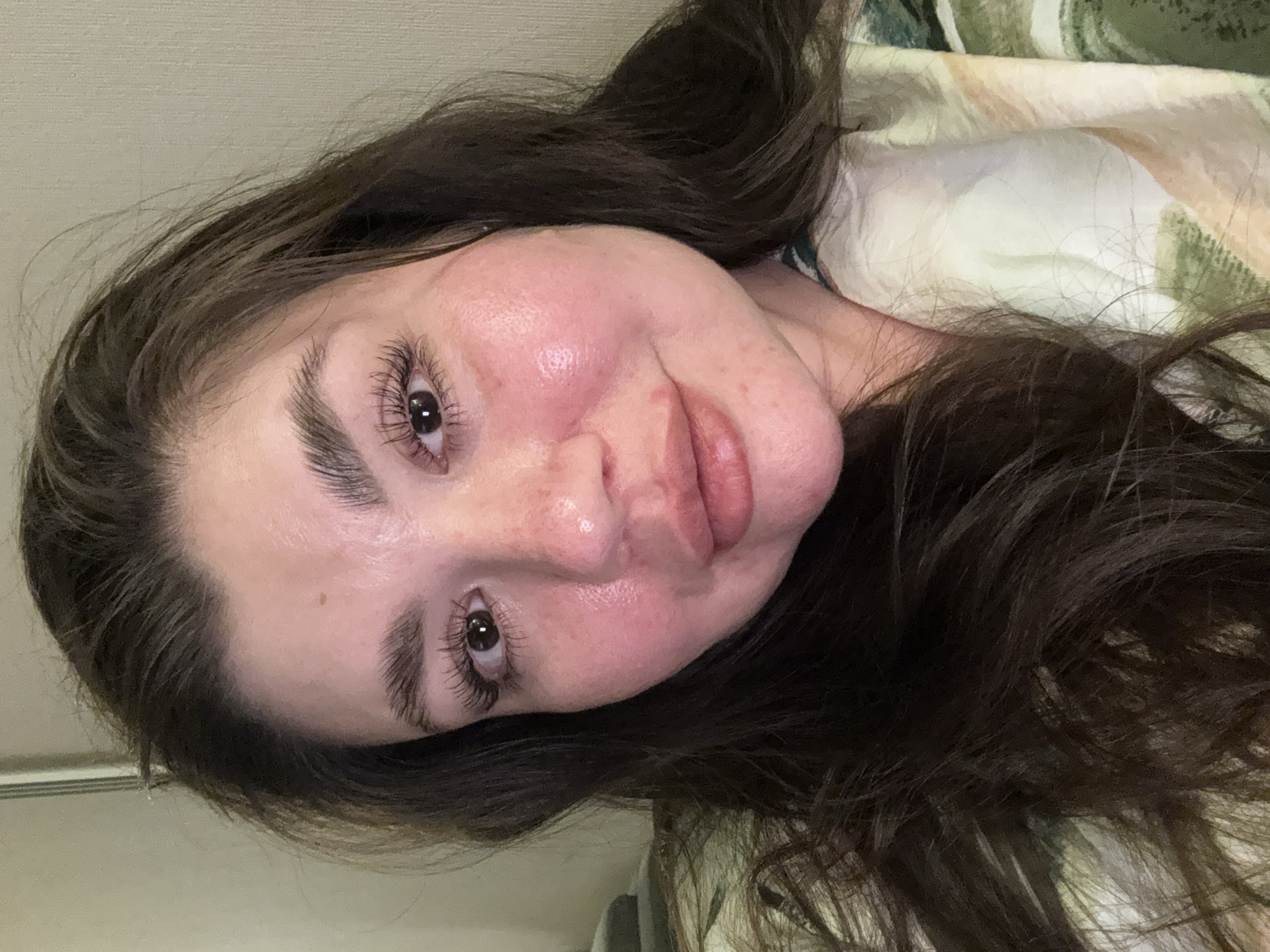 A close-up, slightly angled shot of a woman with long, dark hair. Her skin appears flushed or red, and she has visible pores. Her eyebrows are dark and well-defined. She has brown eyes with long eyelashes. The background includes a beige wall and part of a patterned pillow or blanket. The location is not discernable from the image and cannot be determined to be Votkinsk, Russia.