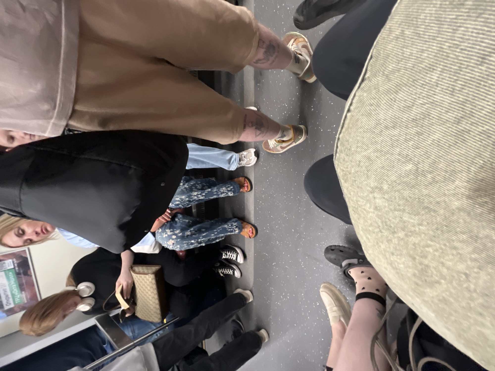 This is a waist-up, eye-level shot taken inside a crowded Saint Petersburg Metro train carriage. Several individuals are seated and standing, their legs and lower torsos filling the frame. In the foreground, a person's lap and thighs are visible, wearing tan shorts and socked feet with visible tattoos on their calves. To the left, another person's legs are visible, one wearing a light-colored loafer and the other wearing black socks with white hearts and a dark Croc shoe. Further back, a woman with blonde hair is seated, wearing a blue shirt and patterned pants, with her hands clasped. Next to her, a woman with reddish-brown hair wearing headphones is reading a book and holding a patterned purse. The floor of the train is a speckled grey surface, and overhead, a portion of a train advertisement is visible. The overall impression is one of a typical, busy commute.