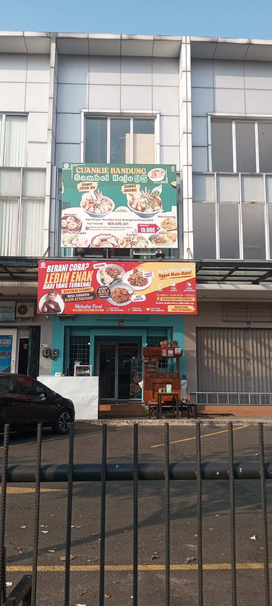The image shows the exterior of a food establishment in Lambangjaya, Indonesia. Two prominent banners are displayed. The upper banner, green and white, advertises "CUANKIE BANDUNG Sambel Hejo 95" and lists various food items like Dimsum, Es Pisang Ijo, Pempek, Cuanki, Bakmi, and Nasi Goreng. It also provides a phone number (0878-8390-2471) and mentions catering services. The lower banner, red and yellow, features a cartoonish figure of a man giving a thumbs-up and promotes "BERANI COBA? LEBIH ENAK DARI YANG TERKENAL!!" with text indicating "BUKTIKAN SENDIRI! RASA JUARANYA BIKIN KE TAGIHAN!". It also advertises "MEHABA FOOD" and lists Bakmi, Bakmi Goreng, and Nasi Goreng. The establishment appears to be a small shop with a blue facade and a glass door. A wooden food cart is positioned outside the entrance. A dark-colored car is parked in front of the building. The scene is set against a backdrop of a multi-story building with a modern facade. The environment suggests a commercial area with clear blue sky overhead.