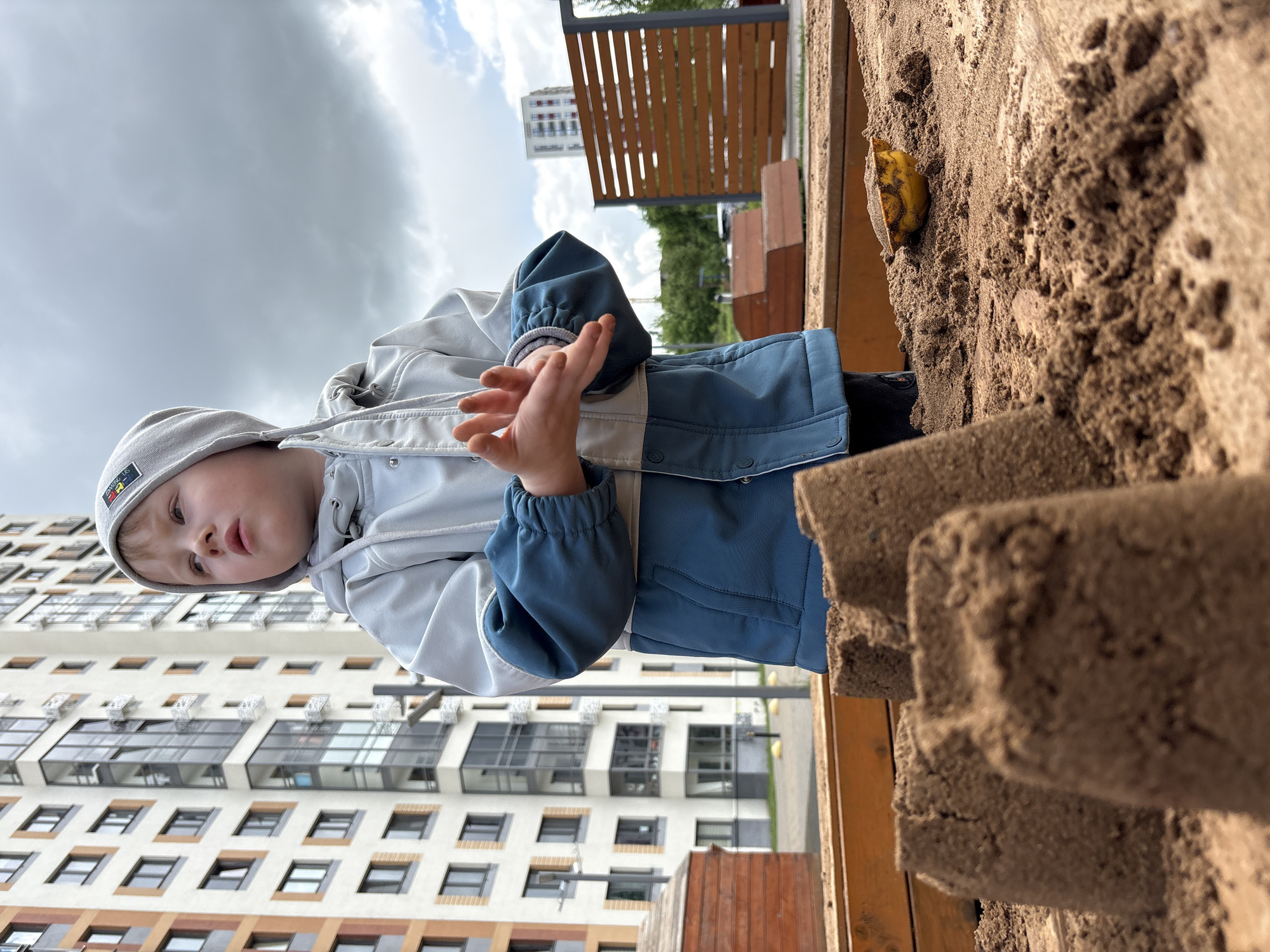 A young child is playing in a sandbox. The child is wearing a light gray and blue jacket with a matching hat. The sandbox is made of wood and filled with sand. Behind the child is a modern apartment building with many windows. In the background, a park and another building are visible under a cloudy sky. The image was taken in Izhevsk, Russia.