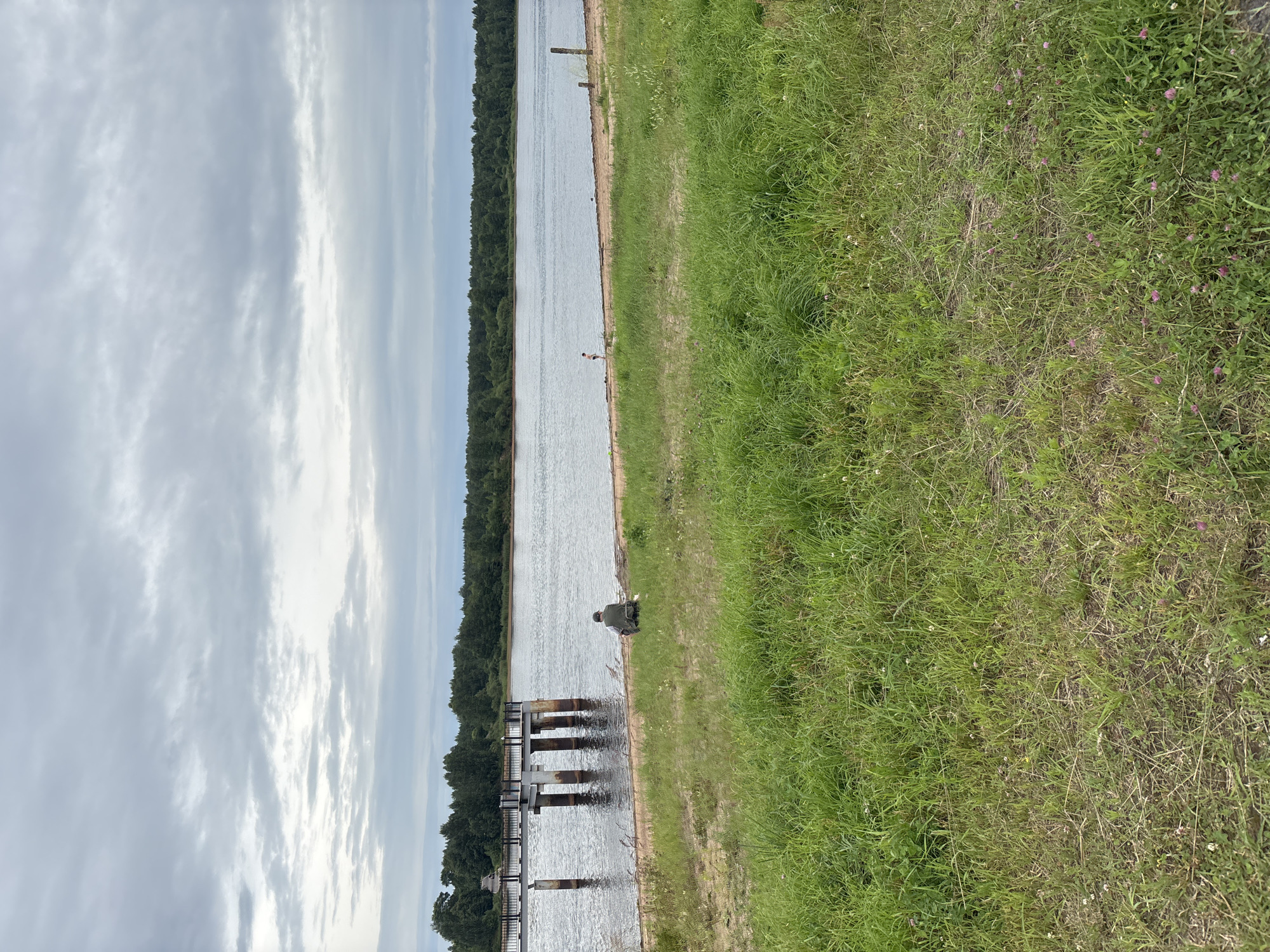 A person is sitting on the grass by the river in Nevskaya Dubrovka, Russia, looking out at the water. The sky is overcast, and the clouds are gray and heavy. A pier juts out into the river on the left side of the image. The shore is lined with trees, and there is a sandy beach in the foreground. The grass is green and lush, with a few small flowers scattered throughout. The overall mood of the image is one of peace and tranquility.