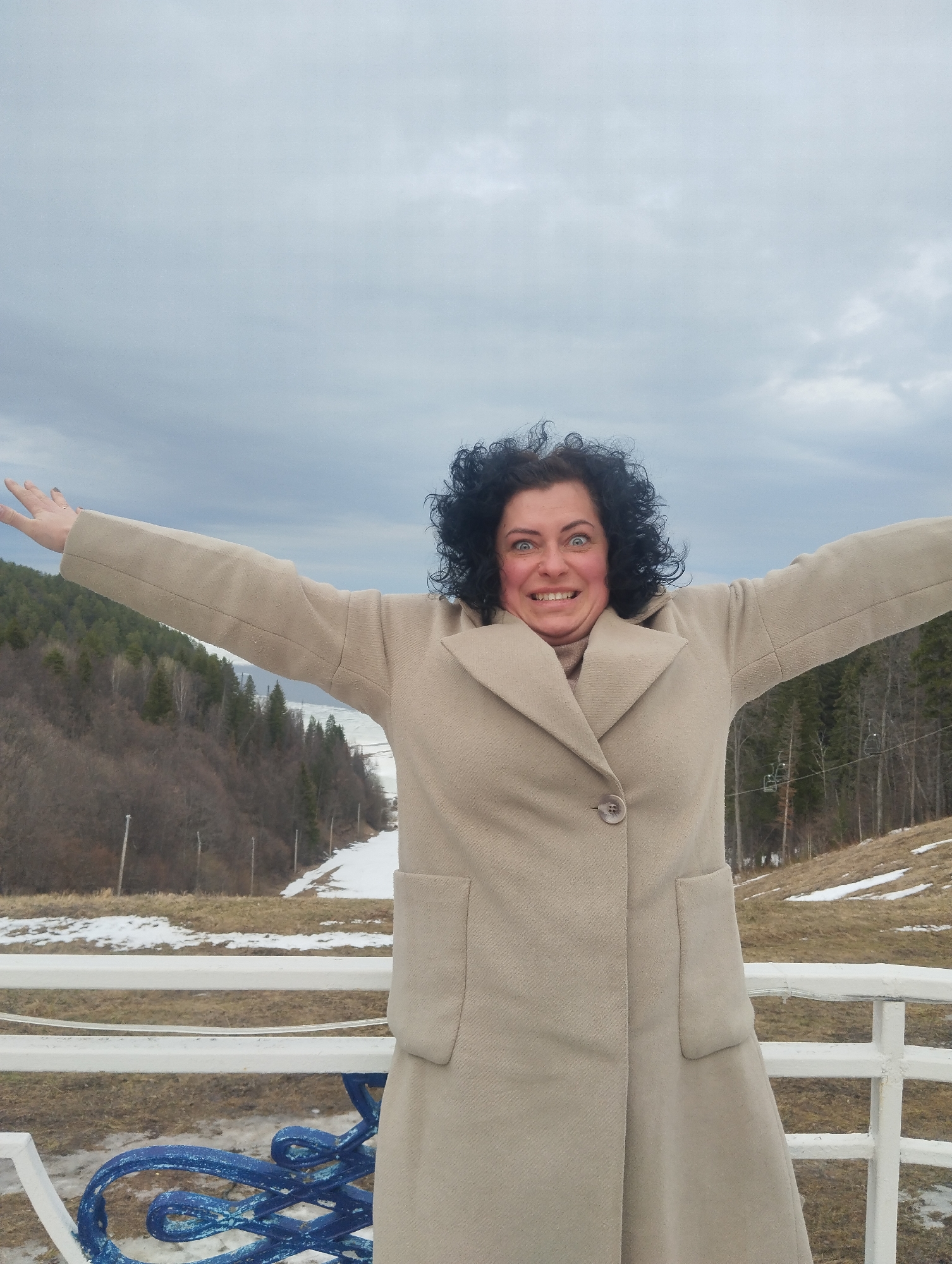 A medium shot depicts a fair-skinned woman with curly black hair and bright blue eyes, broadly smiling with her arms outstretched, positioned in front of a wintry landscape. The woman is wearing a light beige, knee-length coat with a single visible button and large rectangular pockets. She is standing behind a white metal railing, partially obscuring a blue ornamental metal structure at the bottom of the frame.
The background reveals a scene in what appears to be Galevo, Russia, during the colder months. Patches of snow dot the grassy hillsides, leading up to a forested area comprised of coniferous trees. A strip of snow runs down the center of the hill, suggesting a ski slope. The sky is overcast with a dense cloud cover, contributing to the wintery atmosphere. The overall setting is a mountainous area with a cold climate.