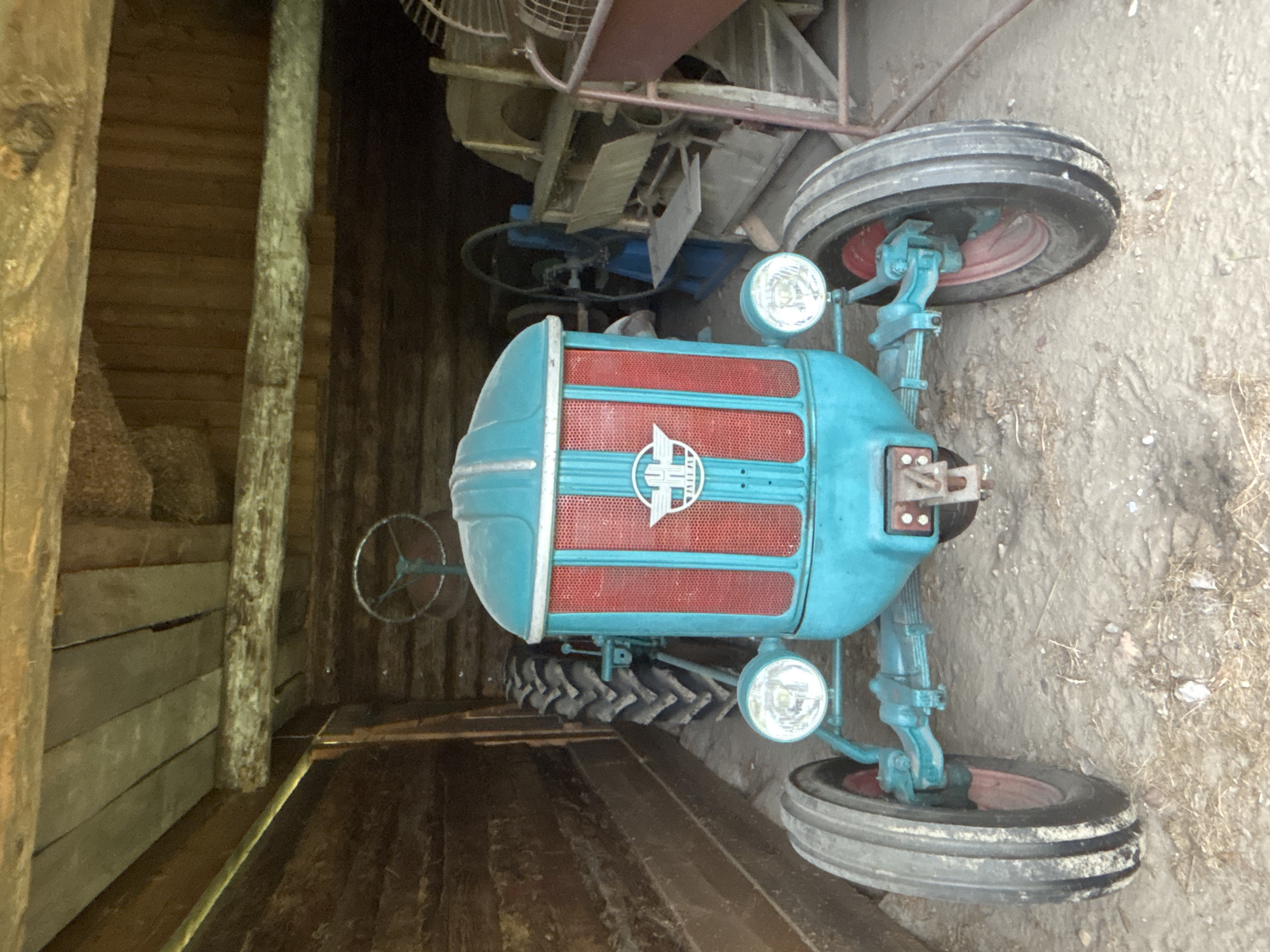 In a rustic wooden shed in Sielachowskie, Poland, a turquoise and red vintage tractor is parked. The tractor's front grill is a prominent red with vertical slats, and it features a circular emblem with a winged symbol. Two round headlights are mounted on the front axle. The tractor has large, treaded tires on the rear and smaller ones on the front, with red rims visible on the front wheels. The shed's interior is constructed from rough-hewn wooden logs and planks. Behind the tractor, other agricultural machinery is visible, including a fan and what appears to be a threshing machine. The ground is covered with dirt and scattered debris. No people are present in the image.