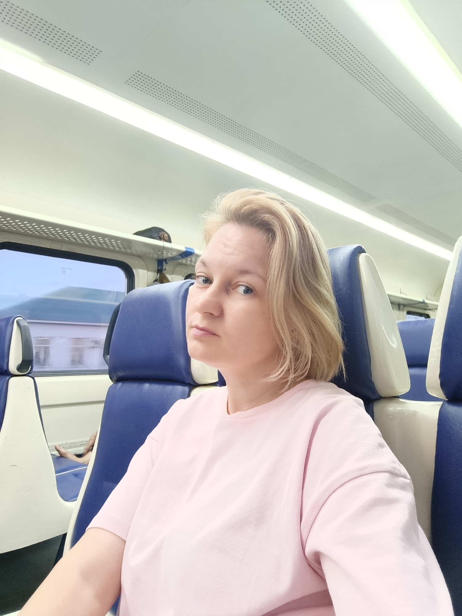 A woman with blonde hair is taking a selfie on a train. She is wearing a pink t-shirt and sitting in a blue and white seat. Through the window, a building can be seen. The train car appears to be modern with white walls and fluorescent lighting. The image was taken in the city of Shemodan, Russia.