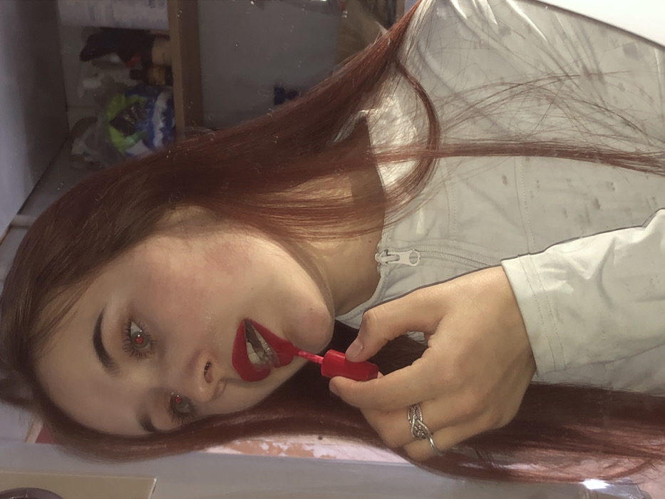 A fair-skinned young woman with auburn hair is applying red lipstick while looking into a mirror. She is wearing a light-colored jacket with a zipper. Her hazel eyes are visible, and she is wearing a ring on her right hand. A shelf with items is visible in the blurred background, reflected in the mirror. The image has a slightly desaturated and slightly blurry aesthetic.