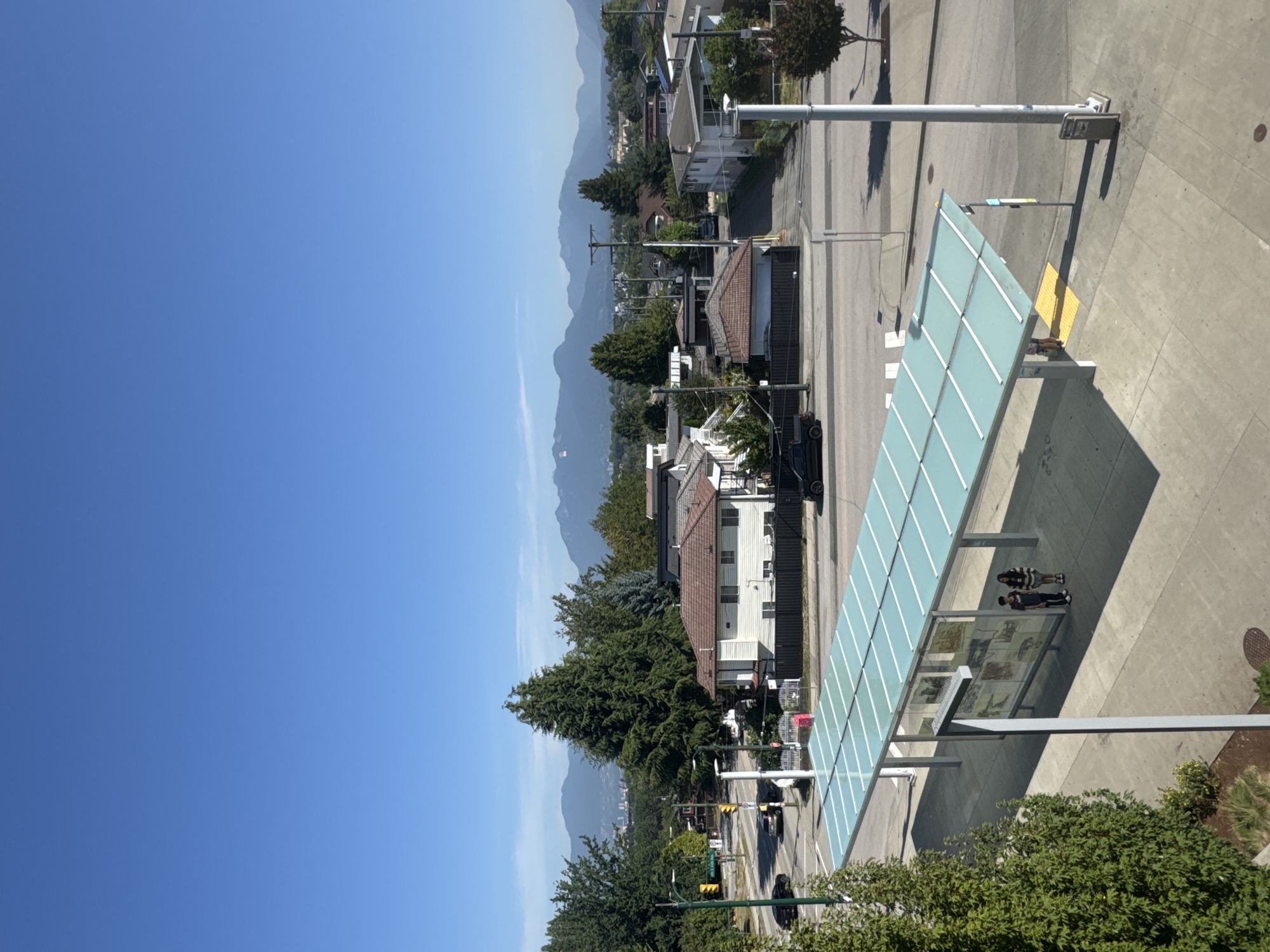 A high-angle outdoor shot captures a suburban street scene in Vancouver, Canada on a clear, sunny day. The foreground features a bus shelter with a translucent blue roof supported by gray metal poles. Two individuals, a male and a female, stand beneath the shelter, facing towards each other. The sidewalk extends to the right of the shelter, where a trash receptacle and a light pole are visible. Beyond the sidewalk, a street with a crosswalk is present, with two vehicles, a black sedan and a black SUV, driving on it. Residential houses with brown roofs and white siding line the opposite side of the street, interspersed with trees. In the background, a dense forest of green trees stretches across the landscape, leading to a distant mountain range under a clear blue sky.