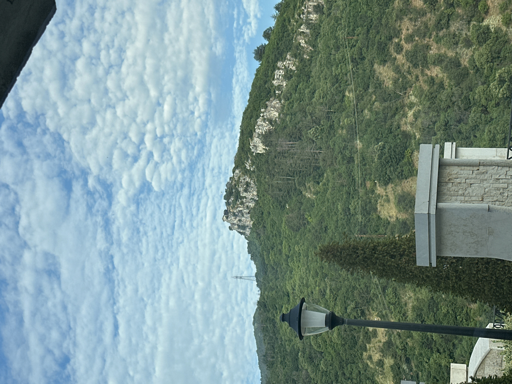 This image depicts a mountainous landscape in Asenovgrad, Bulgaria, under a daytime sky characterized by numerous scattered, small white cumulus clouds against a bright blue background. A large, verdant hill dominates the central and background, densely covered with various shades of green deciduous and coniferous trees. Prominent greyish-white rock formations are visible along the upper slopes of the hill, particularly on the right side. At the highest rocky peak towards the right, a small, indistinct man-made structure, possibly historical ruins, is situated. To the left, on a slightly lower peak, a tall, grey, lattice-structure electrical transmission tower stands out against the sky. Horizontal power lines are faintly visible across the lower sections of the hill. In the foreground, a black, traditional-style street lamp with a clear glass enclosure stands on the left. A tall, slender coniferous tree, dark green in color, is positioned to the right of the street lamp. The bottom right edge of the frame reveals the flat top of a modern, rectangular concrete structure with a textured, stone-clad facade. Another rounded, grey concrete structure is partially visible on the far bottom-left. No individuals or animals are present in the scene, and no specific actions are depicted.