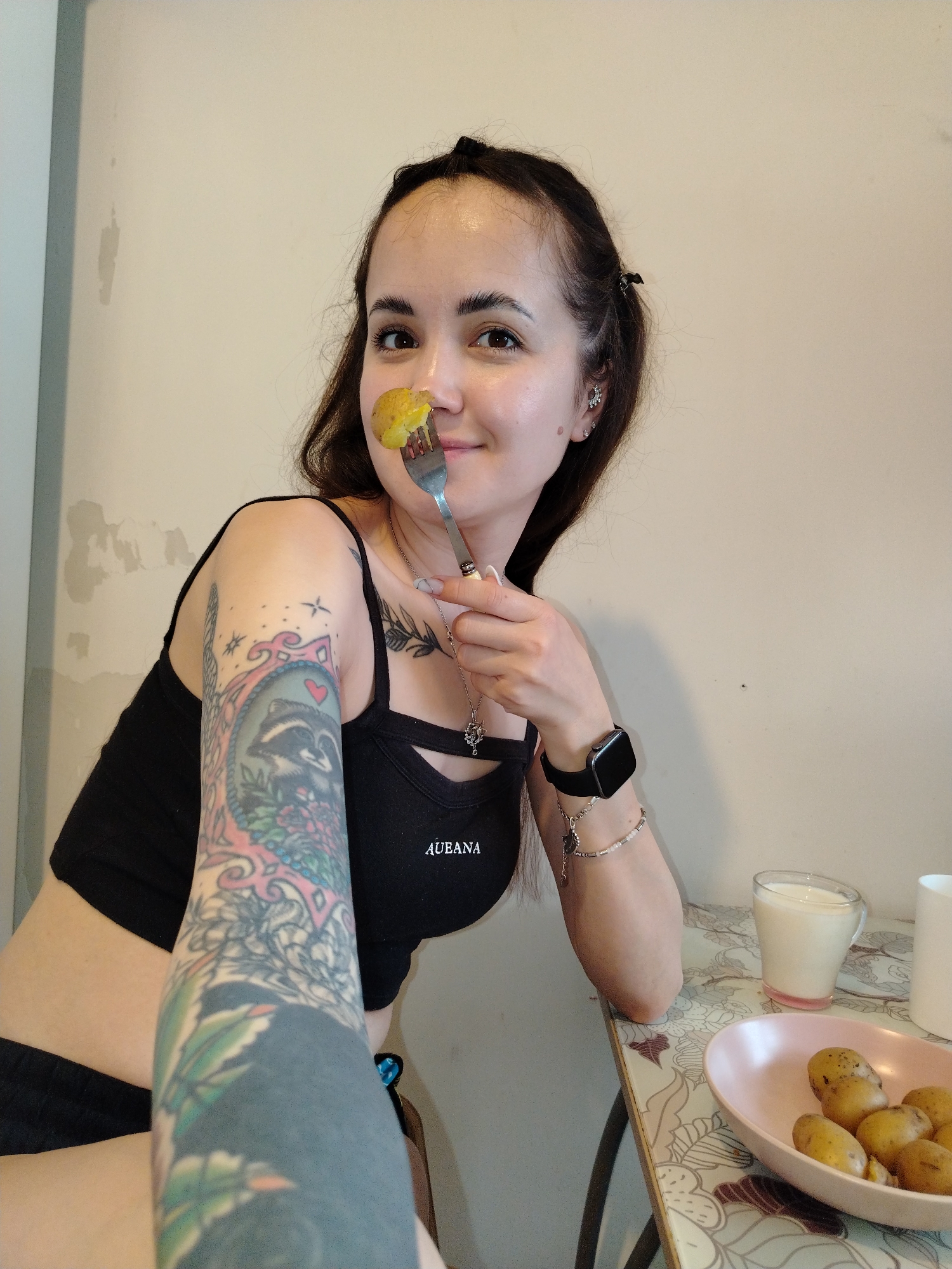 A fair-skinned woman with long, dark hair pulled back from her face is seated at a table and holding a piece of food on a fork in front of her nose. She is smiling and looking directly at the camera. She is wearing a black crop top and dark shorts, a smartwatch on her left wrist, and several bracelets. She has numerous tattoos on her left arm. A shallow, pink dish containing small, golden-yellow potatoes sits on the table in front of her, along with a glass of a light-colored liquid and a white mug. The table has a floral patterned top. The background consists of a plain, light-colored wall with some patchy discoloration. No city or location can be identified from the given image.