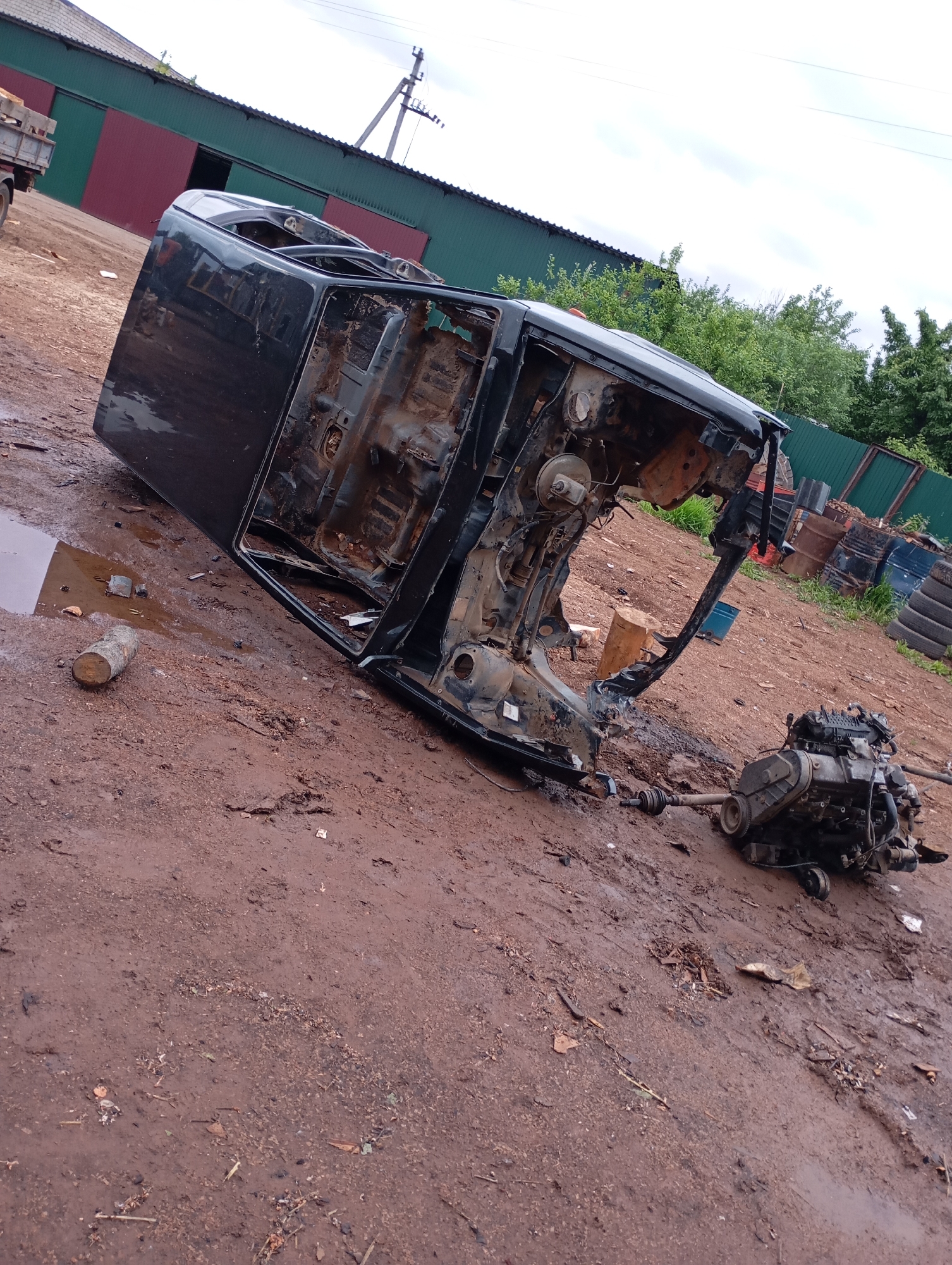 The image displays an outdoor scene in Фоки, Russia, characterized by an unpaved, muddy ground under an overcast sky. In the foreground, a heavily stripped black vehicle chassis, resembling an SUV or truck cab, rests on its driver's side, exposing its rusted underside and internal structure; doors and interior components are absent. Adjacent to the chassis, a complete engine block with an attached transmission lies on the ground, connected to the vehicle frame by a driveshaft component.
To the left of the scene, a large structure with a green roof and segmented green and reddish-brown walls is partially visible, with an open doorway in the reddish-brown section. The rear bed of a light-colored truck, containing wooden items, is parked in front of this building. A utility pole with associated power lines stands further back on the left.
The right side of the background features a dense cluster of green trees and bushes, a green metal fence, and an accumulation of industrial materials including stacked tires and various metal barrels, some blue and some rusted brown. The ground throughout the scene is reddish-brown dirt, saturated with moisture, forming puddles and displaying debris such as a cut log, scattered wood fragments, and general refuse. No individuals are present in the image.