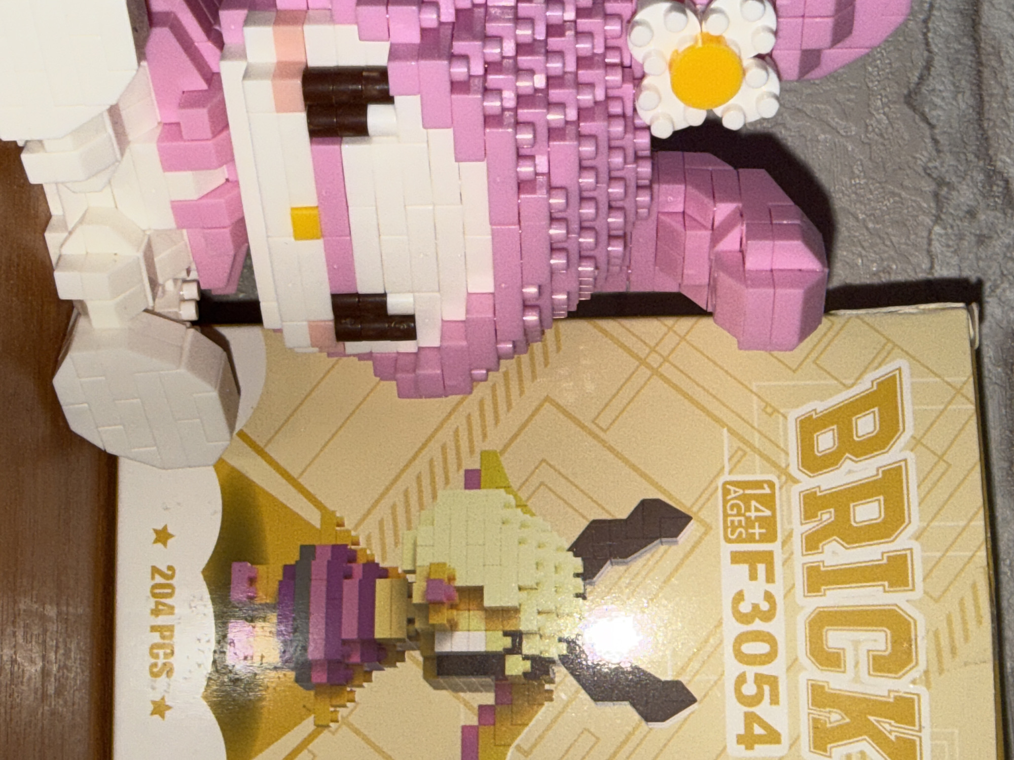The image shows a close-up of a pink and white toy, resembling a character from Sanrio, which appears to be constructed from small interlocking bricks, similar to LEGOs. Next to the toy is the packaging for a "BRICK" toy, with the text "14+ AGES" and "F3054" visible. The box also features an illustration of a character with brown horns and blonde hair, wearing a purple outfit. In the bottom right corner of the box, the text "204 PCS" is present. The background is a textured grey surface, possibly a wall. The toy and box are placed on a brown surface, likely a table or shelf. There is no indication of the location being in Rayevskiy, Russia.